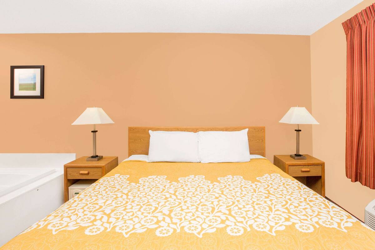 A hotel room with a bed, two bedside tables with lamps, a nightstand, a window with curtains, and a picture on the wall.