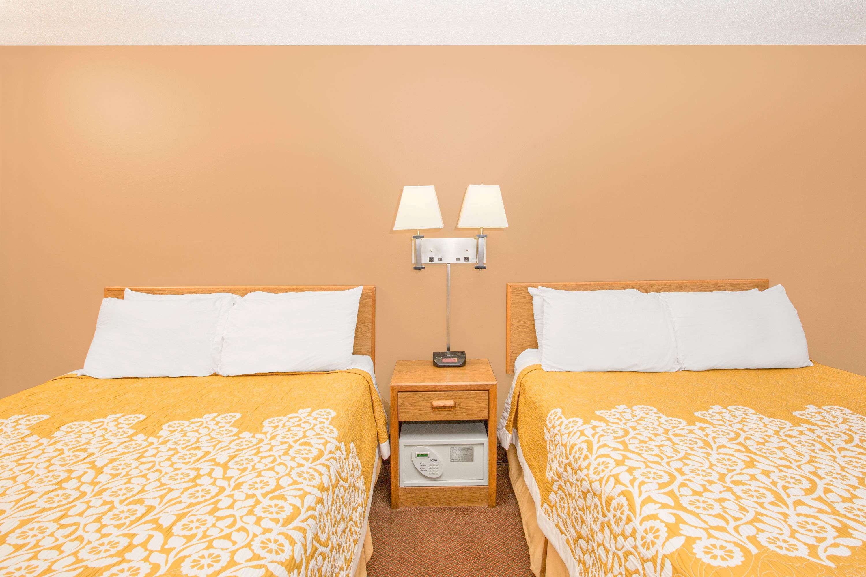 Habitación de hotel con dos camas, cada una con almohadas blancas y una colcha amarilla estampada. Hay una mesita de noche con una lámpara y un panel de control entre las camas.