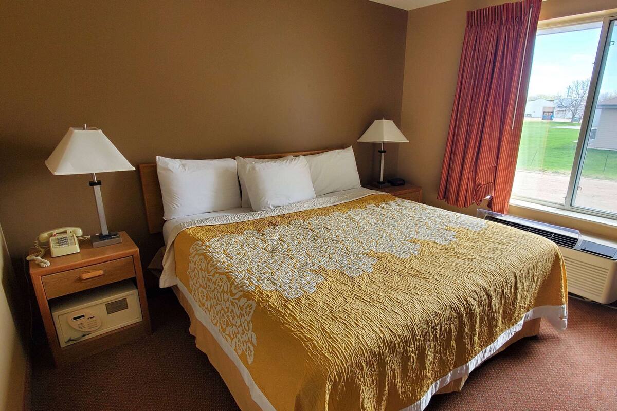 A hotel room with a bed, two bedside lamps, a nightstand with a phone, a window with curtains, and a view of the outside.