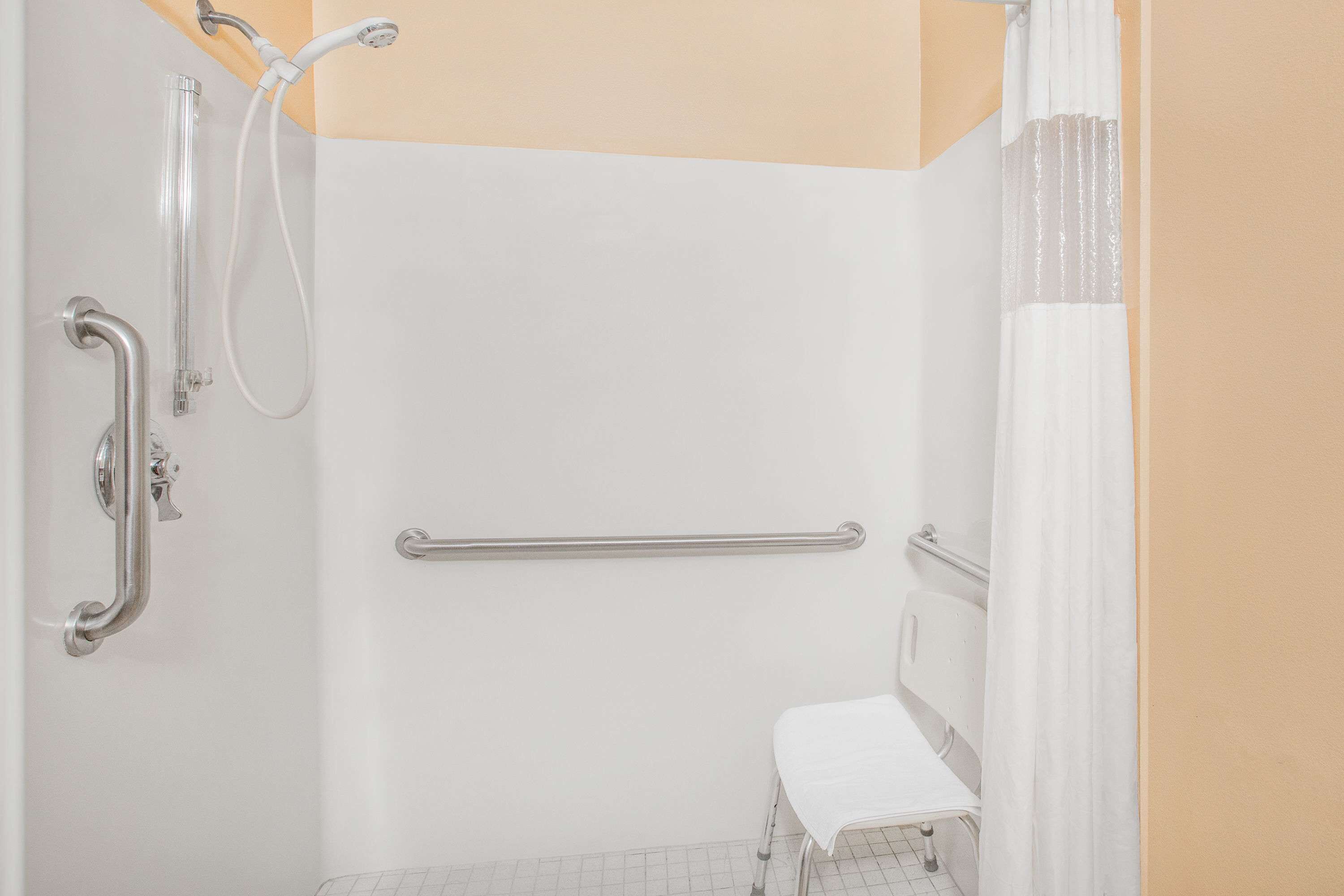 A bathroom with a white bathtub, a shower with a handheld showerhead, a grab bar, and a white bench.