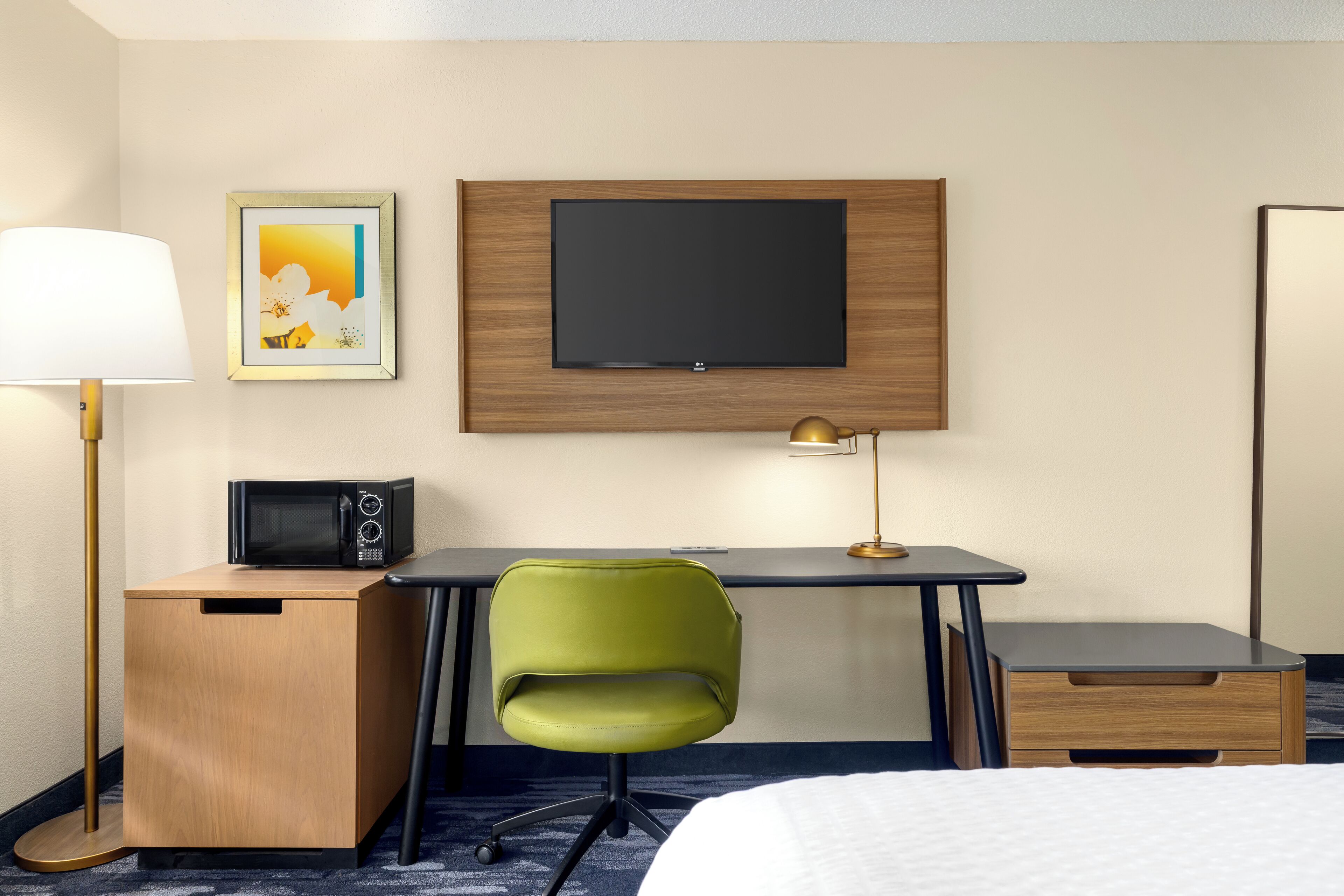 Quarto de hotel com cama, escrivaninha com cadeira, TV de tela plana, micro-ondas, abajur e um quadro na parede.