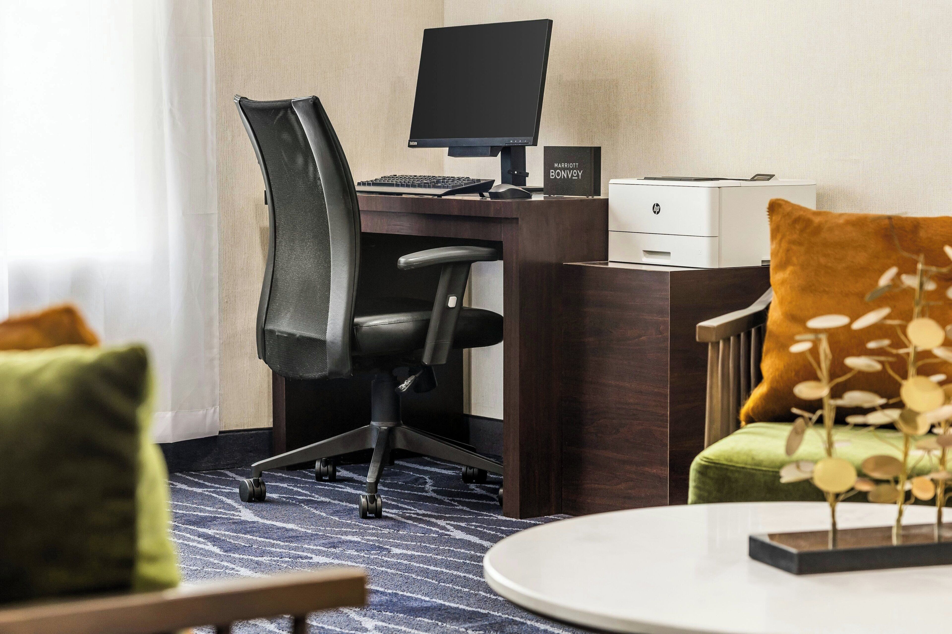 A hotel room with a dark wood desk, a black office chair, a desktop computer, a printer, and a small sign that reads "Marriott Bonvoy".