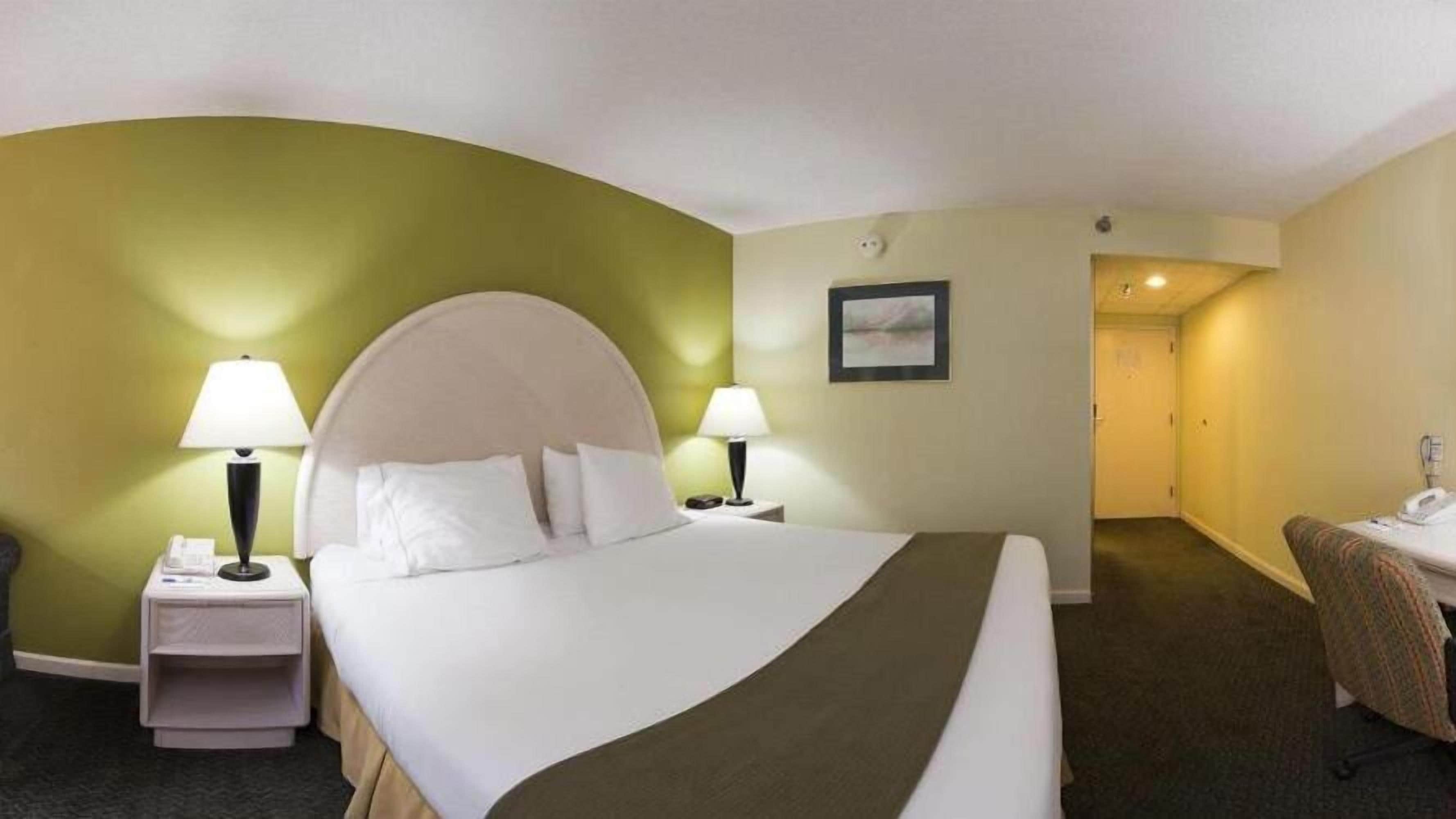 A hotel room with a large bed, two bedside tables with lamps, a small desk with a chair, and a door leading to another room.
