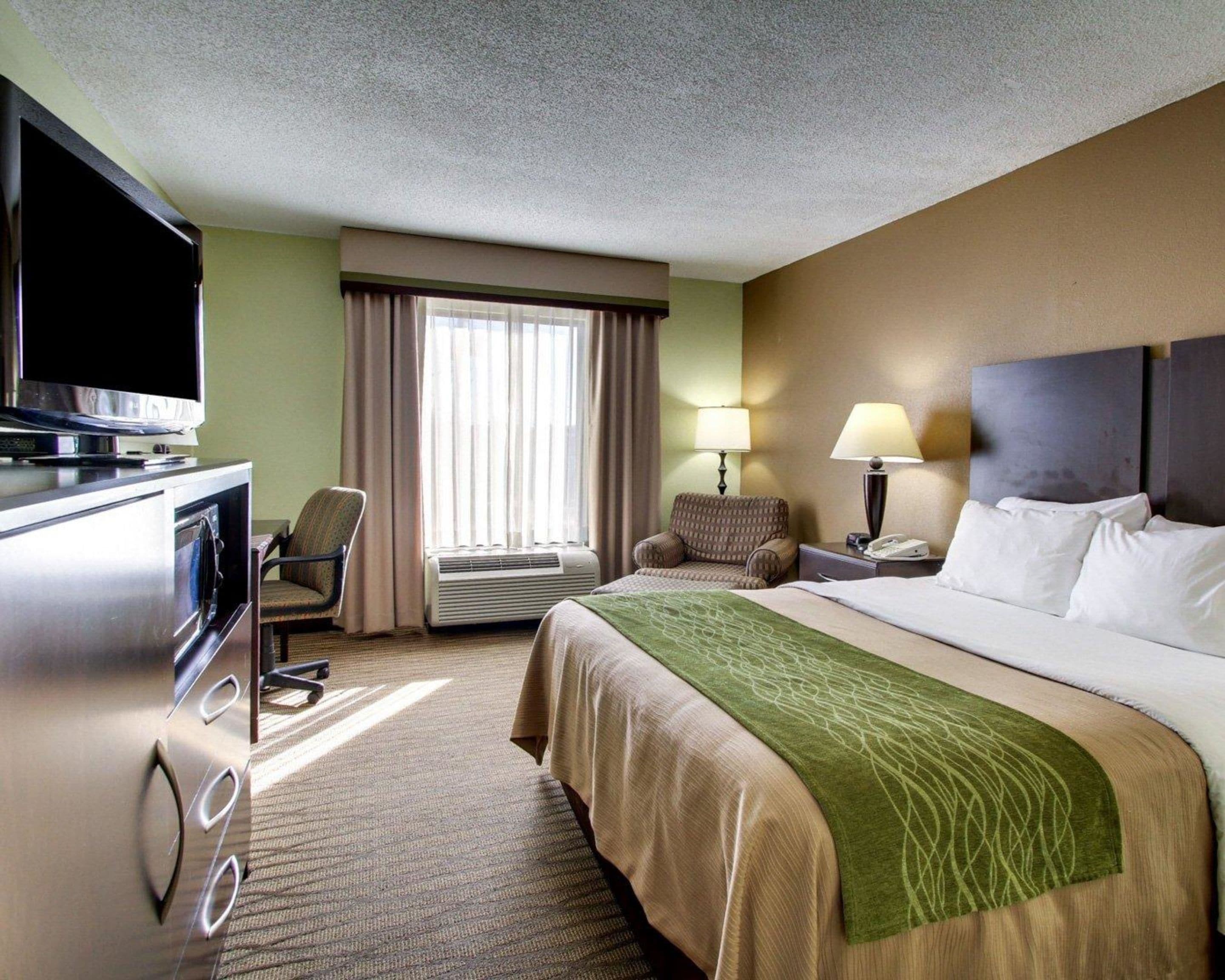 Habitación de hotel con cama, televisión, escritorio, silla, lámpara y ventana con cortinas.