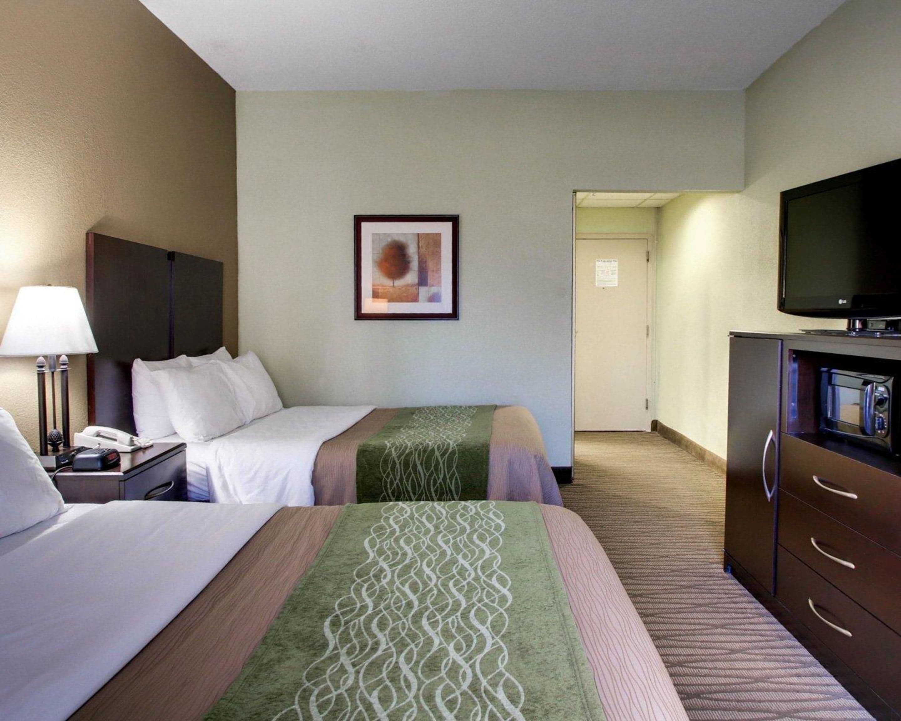 Una habitación de hotel con dos camas, un escritorio con lámpara, un televisor en un mueble y un cuadro en la pared.