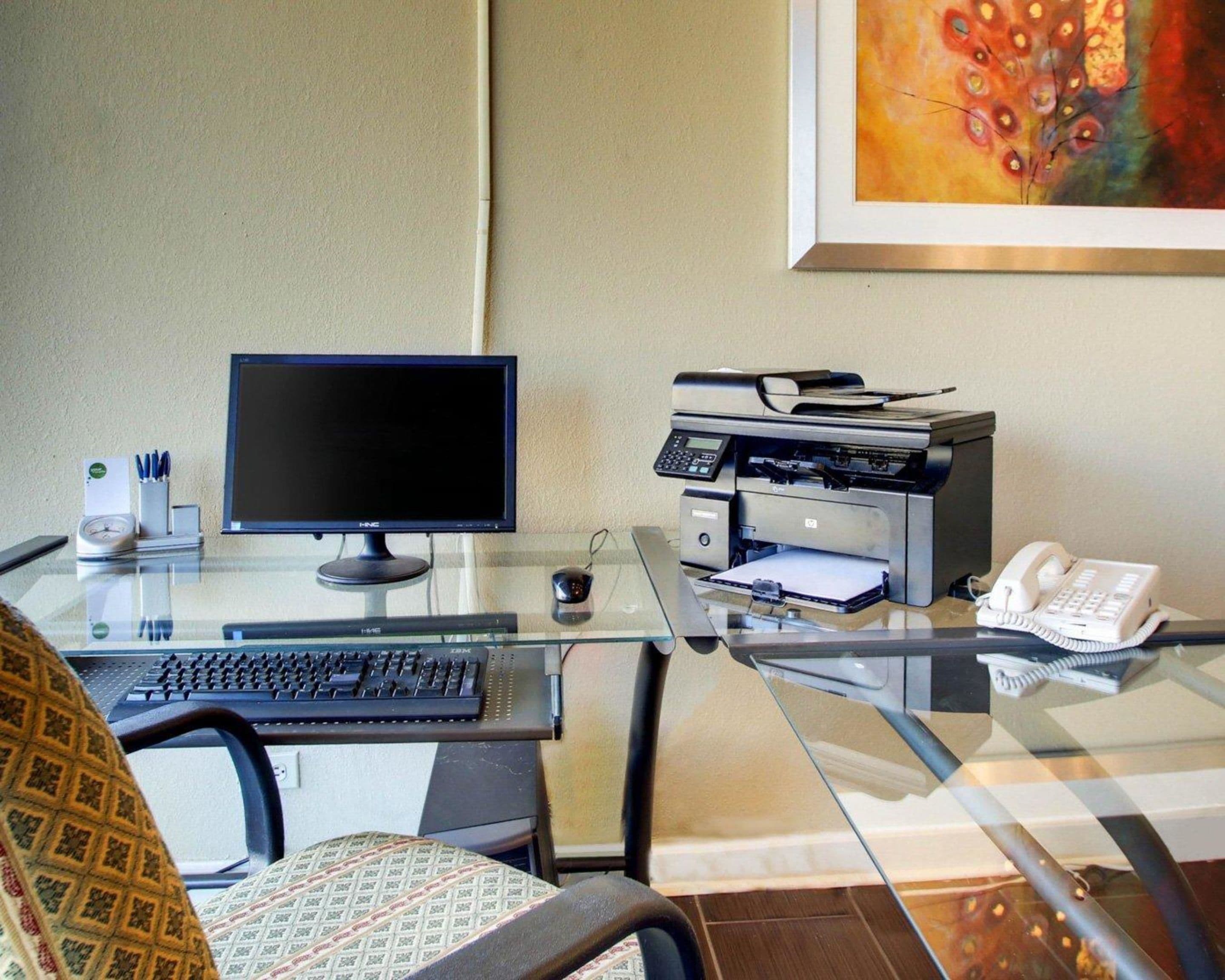 A hotel room with a glass desk, a computer monitor, a printer, a telephone, and a chair.