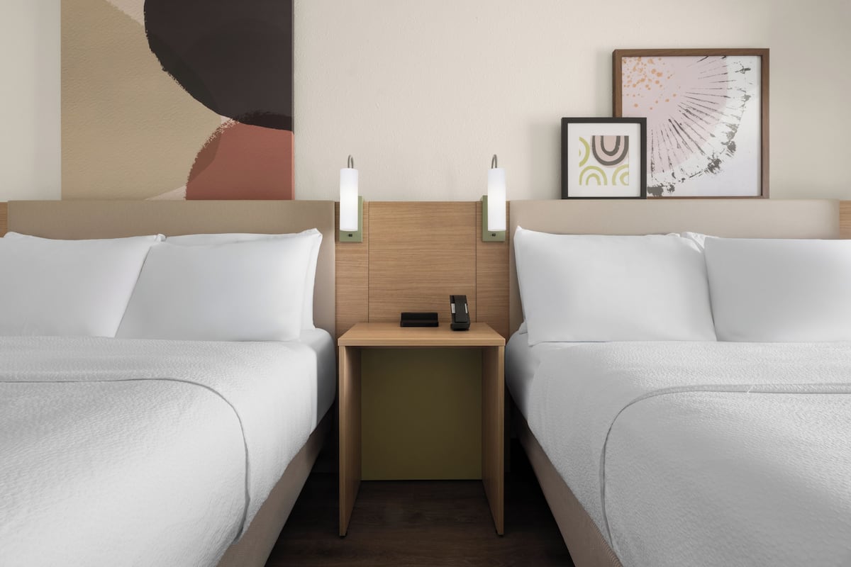 Quarto de hotel com duas camas, mesa de cabeceira com telefone e uma pequena mesa, e arte nas paredes acima da mesa de cabeceira.