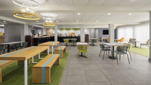 Una cafetería moderna con largas mesas de madera, sillas verdes y detalles en amarillo.