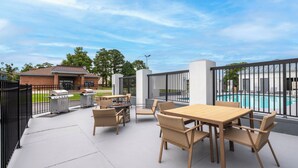 Un patio exterior con mesa de madera y sillas de mimbre, una parrilla y una piscina al fondo.