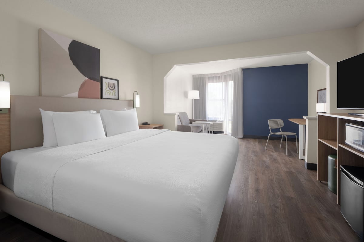 Quarto de hotel com uma cama grande, uma escrivaninha, uma cadeira, uma televisão e uma janela com cortinas.