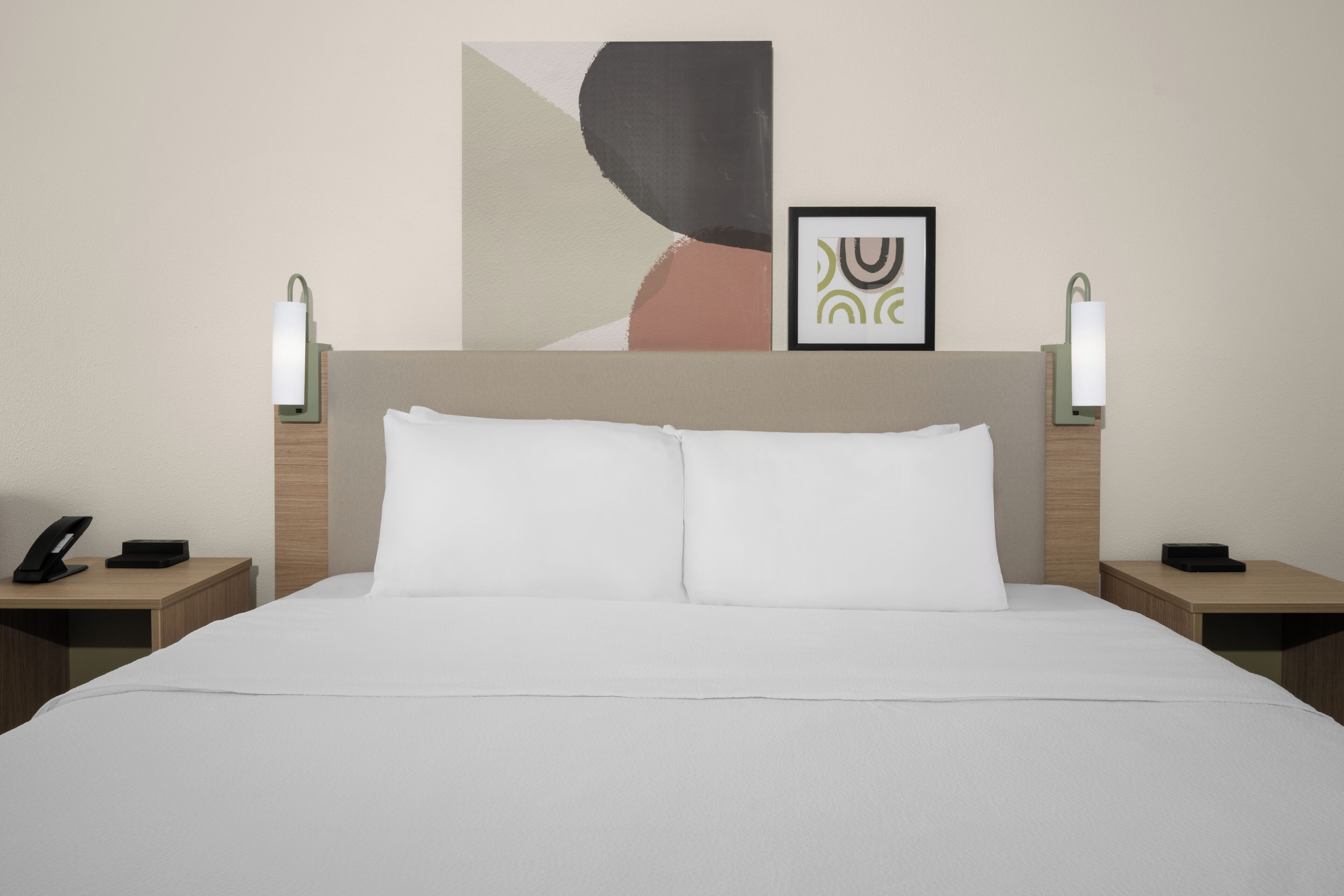 A hotel room with a large bed, two bedside tables, a nightstand, a wall-mounted lamp, and a framed picture.