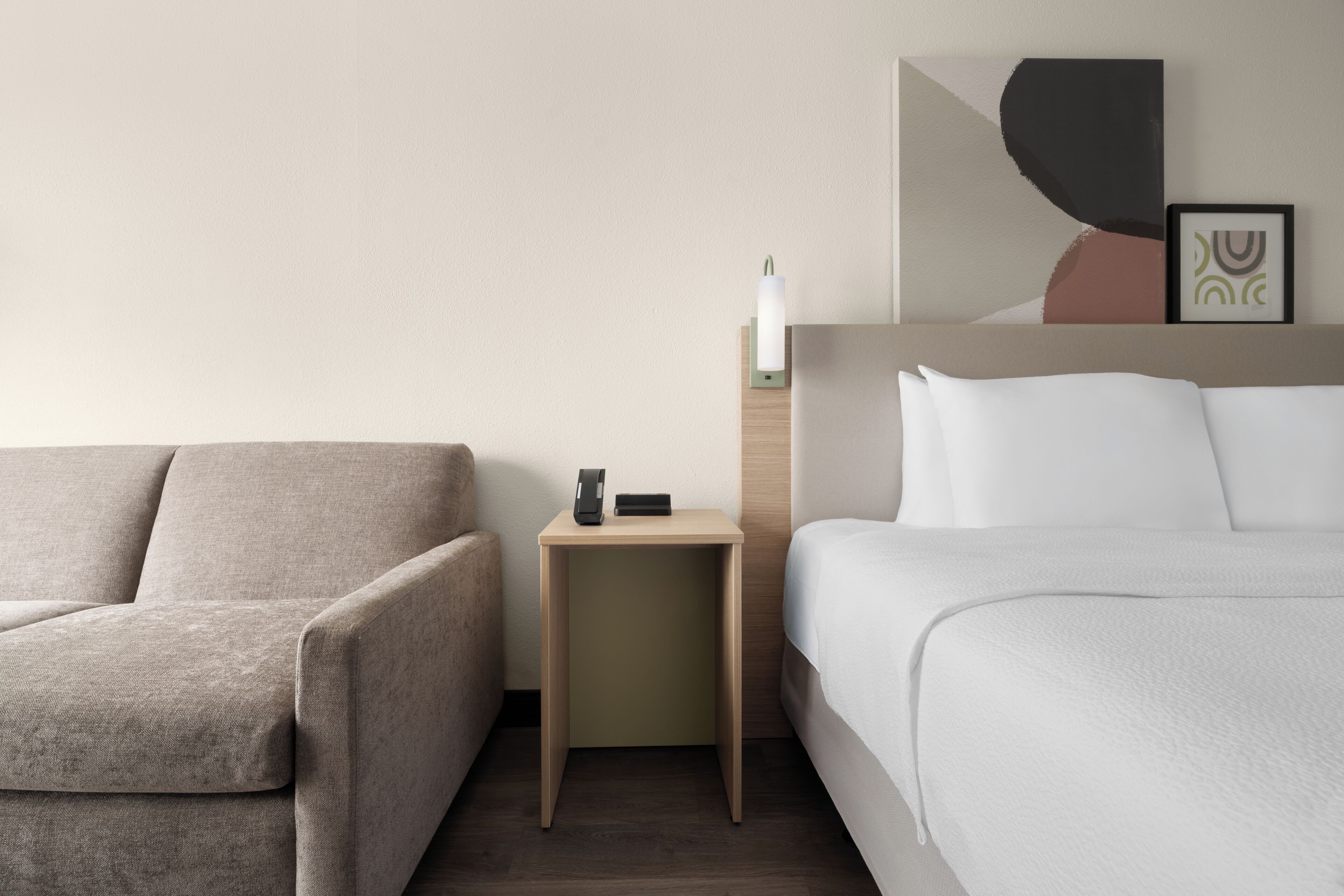 A modern hotel room with a sofa, a bedside table with a lamp, and a bed with white bedding.