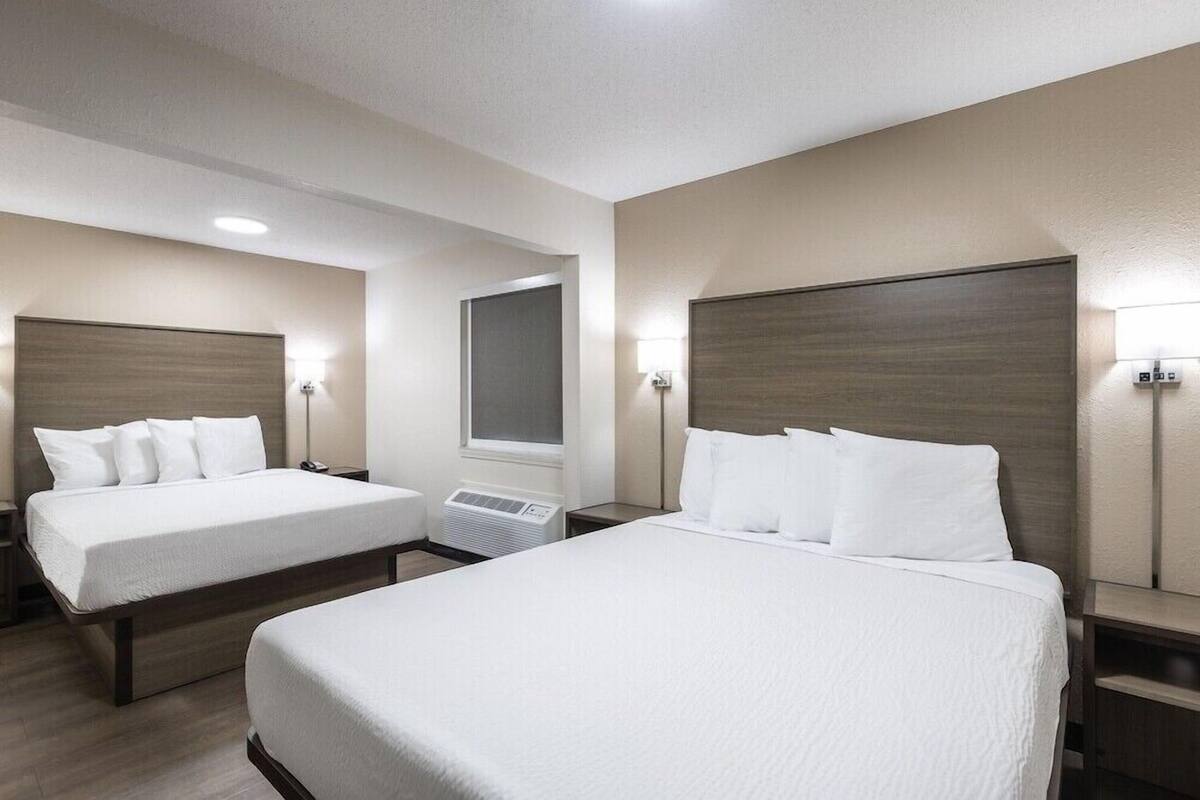 A hotel room with two beds, a window, and a wall-mounted air conditioning unit.