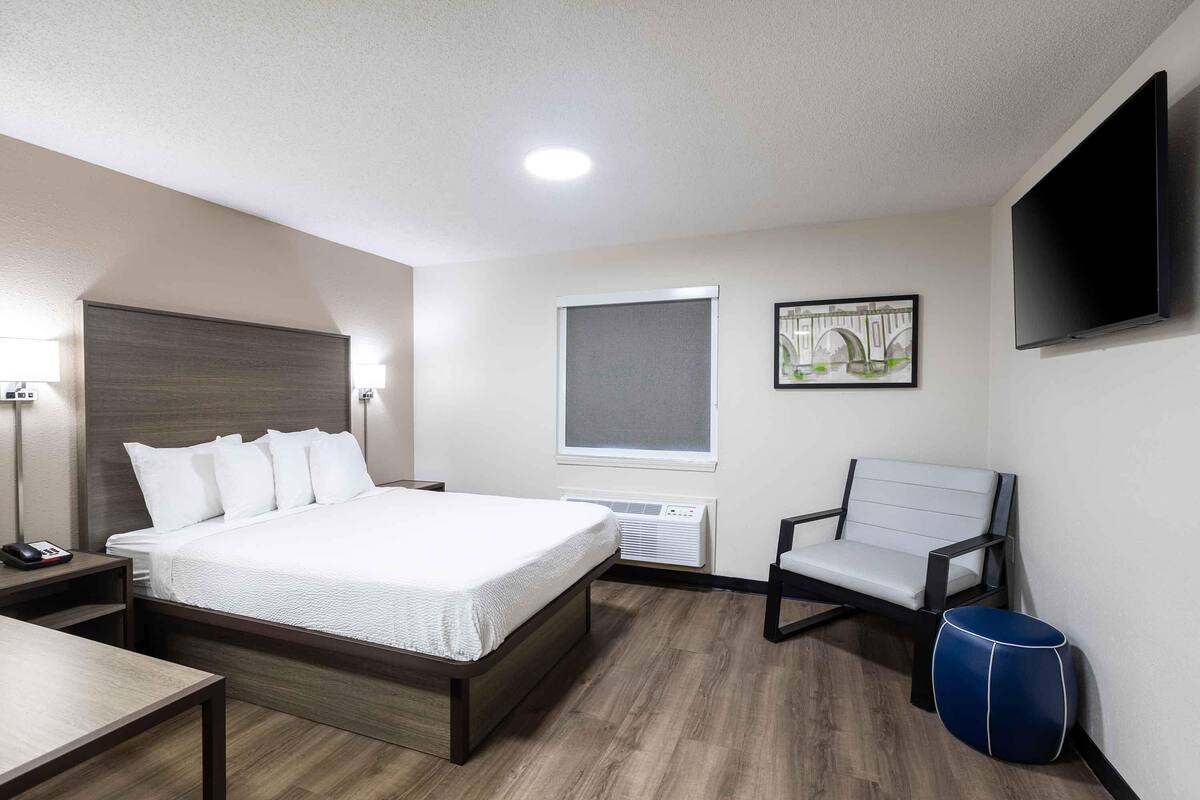 A hotel room with a bed, bedside table, chair, and a television on the wall.