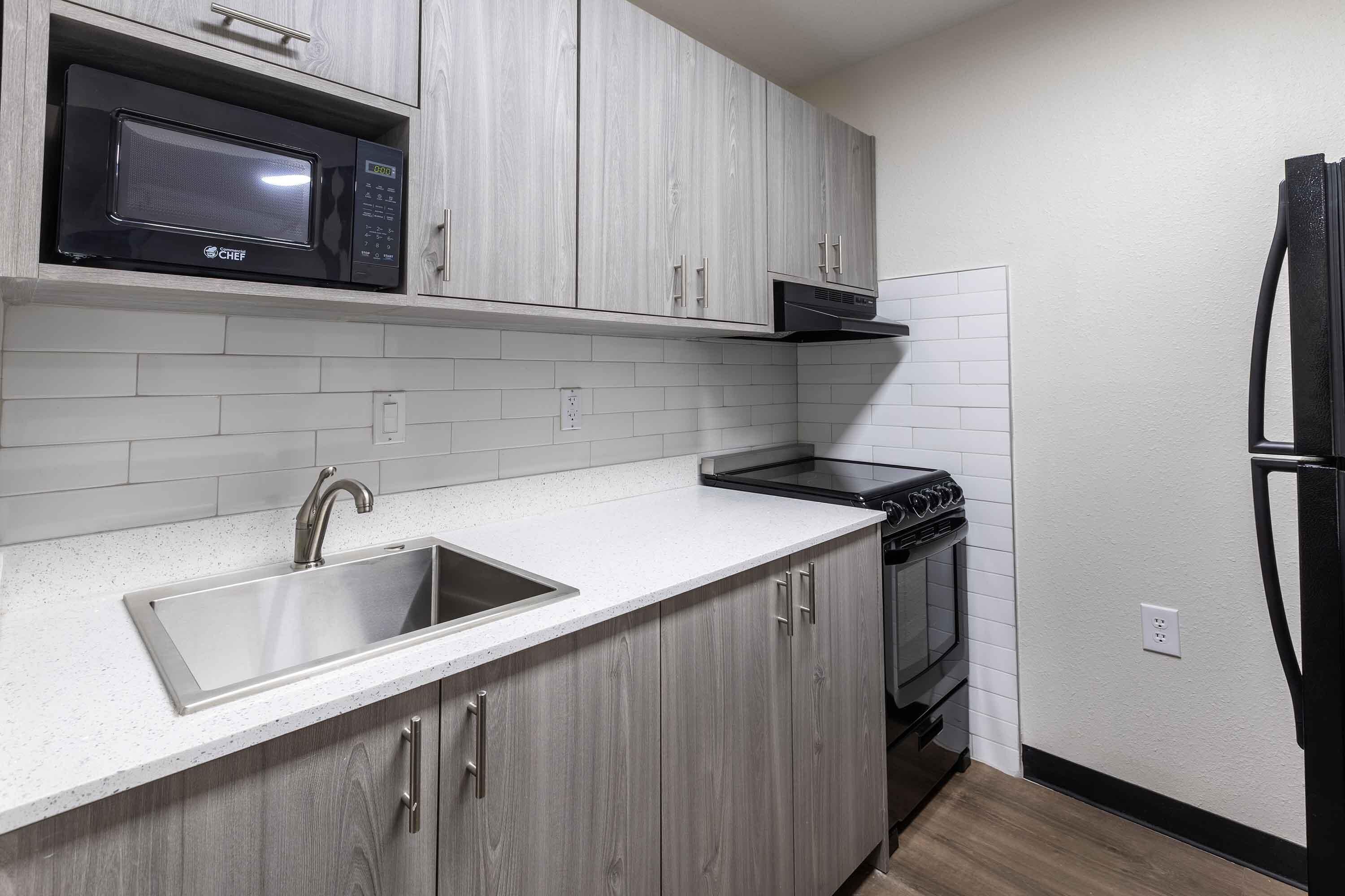 A modern kitchen with grey cabinets, a stainless steel sink, a black microwave, a stove, and a refrigerator.