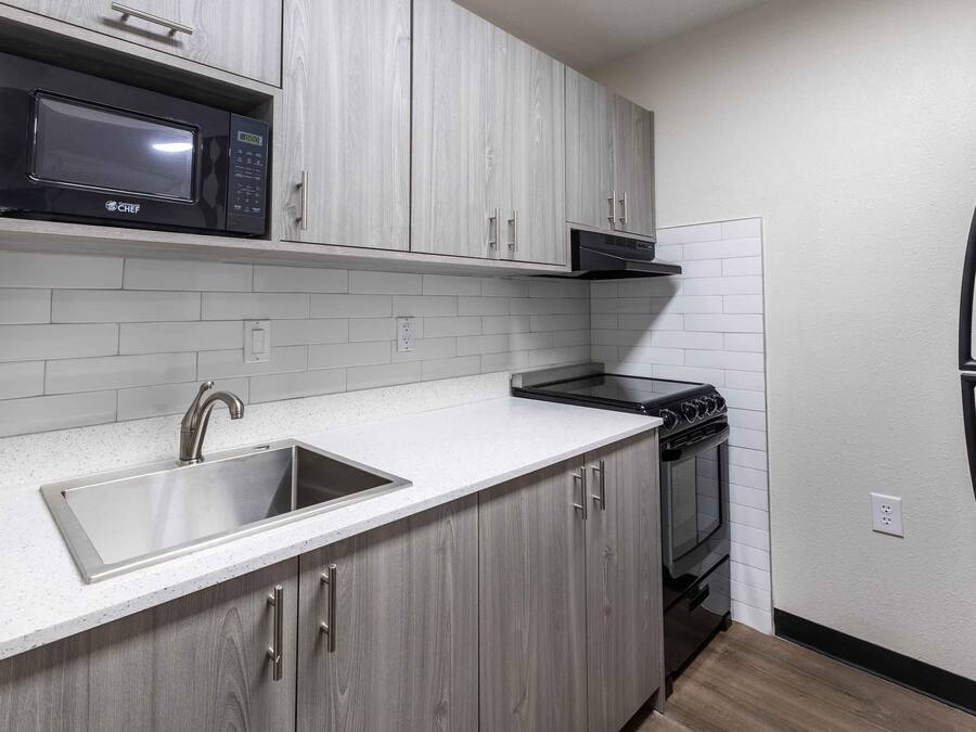 A modern kitchen with grey cabinets, a stainless steel sink, a black microwave, a stove, and a refrigerator.