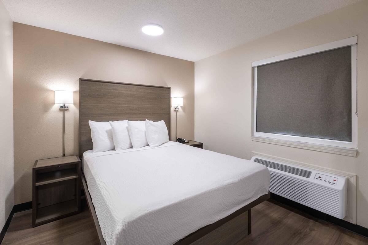 A hotel room with a large bed, two bedside tables, a wall-mounted air conditioner, and a window with a view.