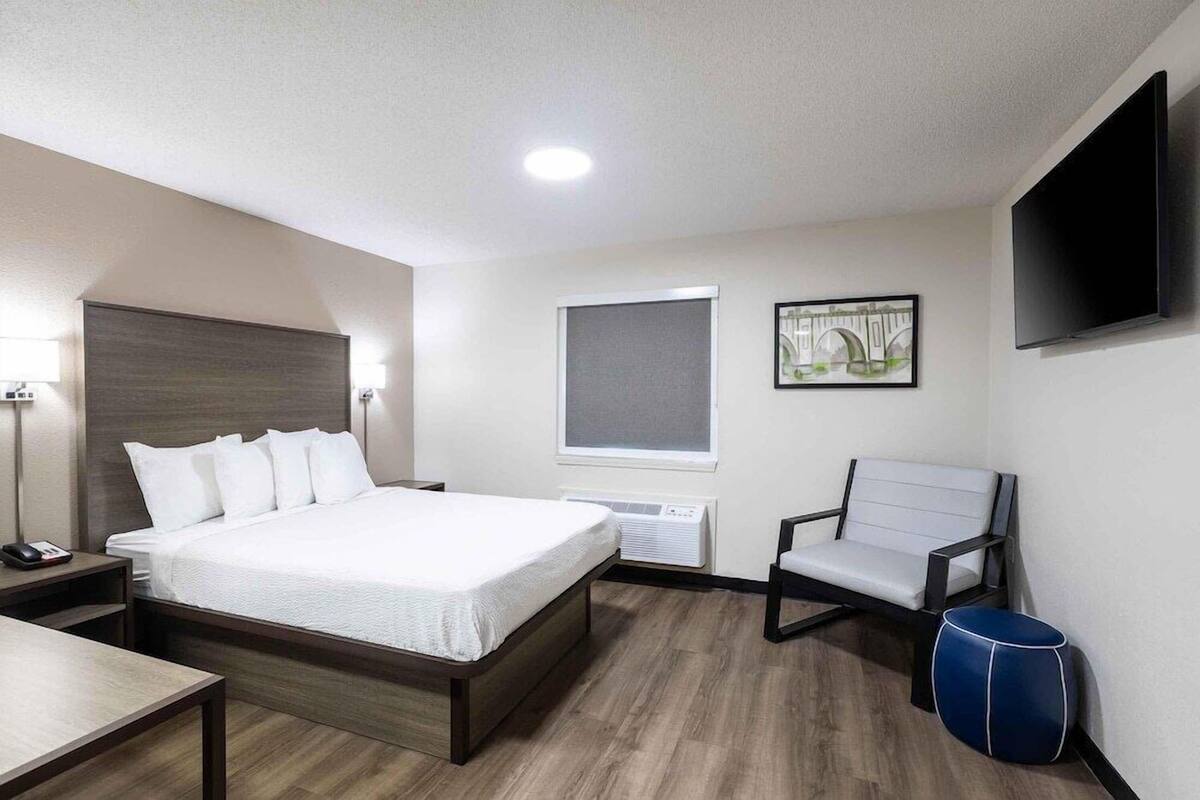 A hotel room with a bed, bedside table, chair, TV, window, and artwork.