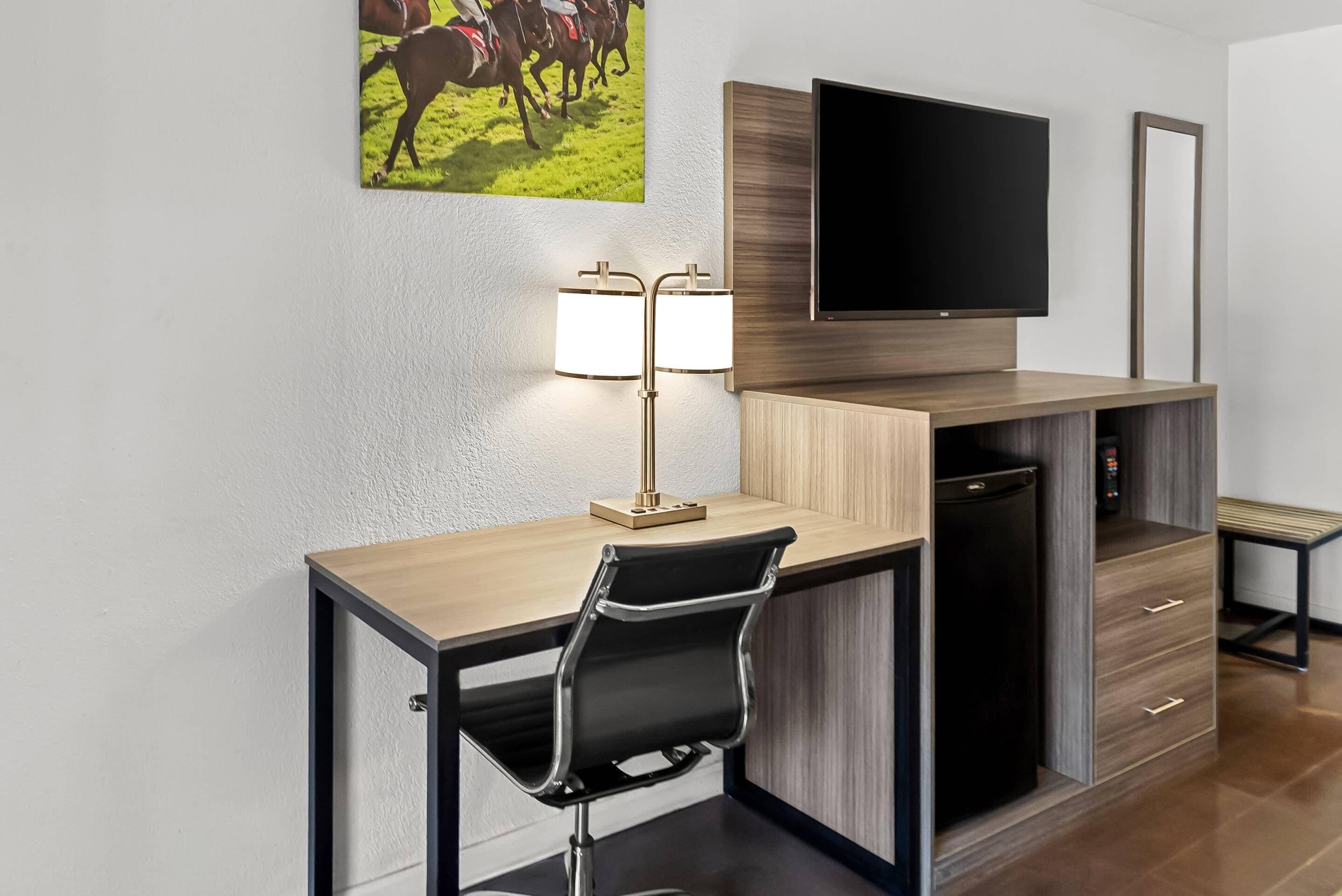 A hotel room with a desk, chair, TV, lamp, and a small refrigerator.
