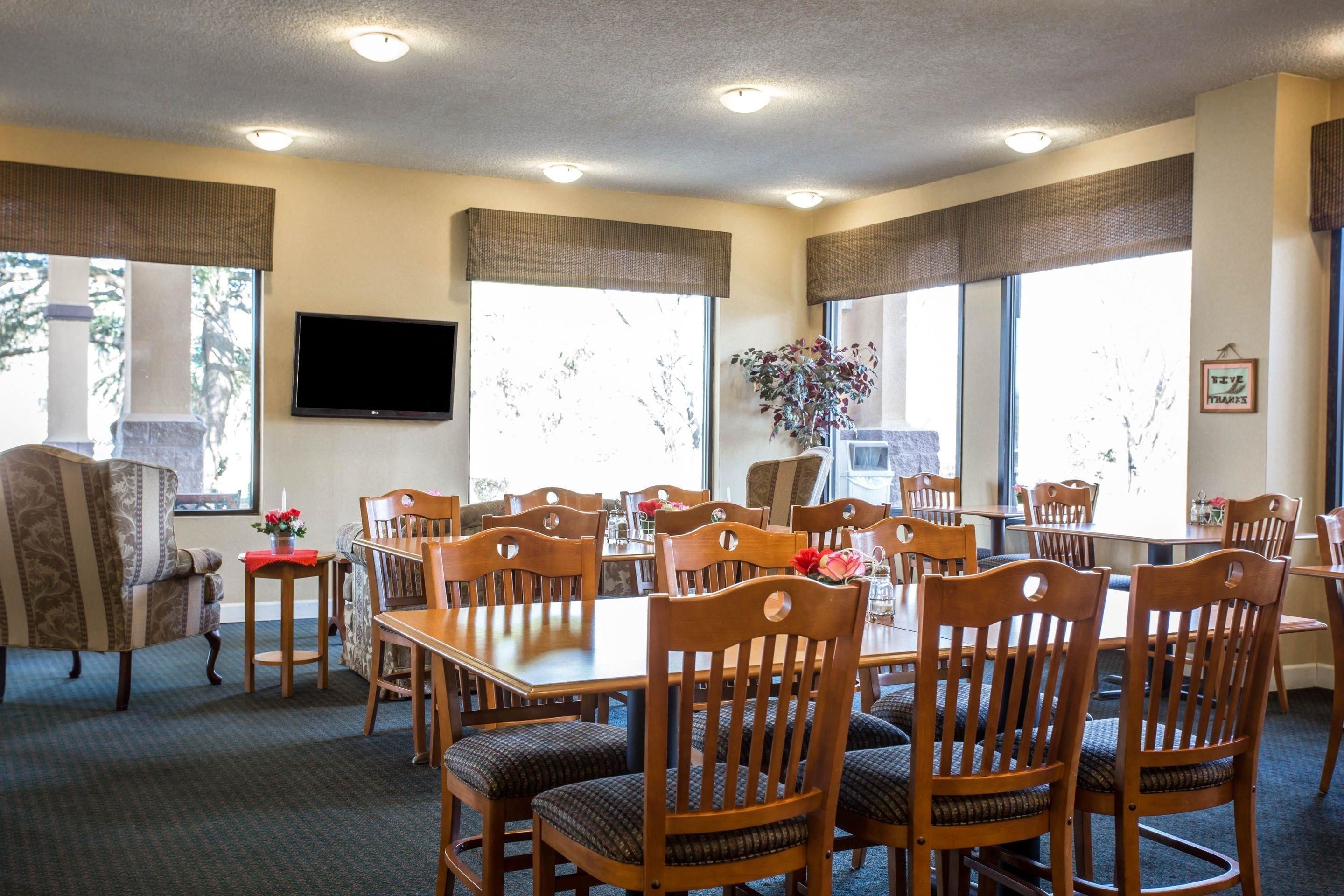 A dining area with multiple wooden tables and chairs, a television mounted on the wall, and large windows with blinds.