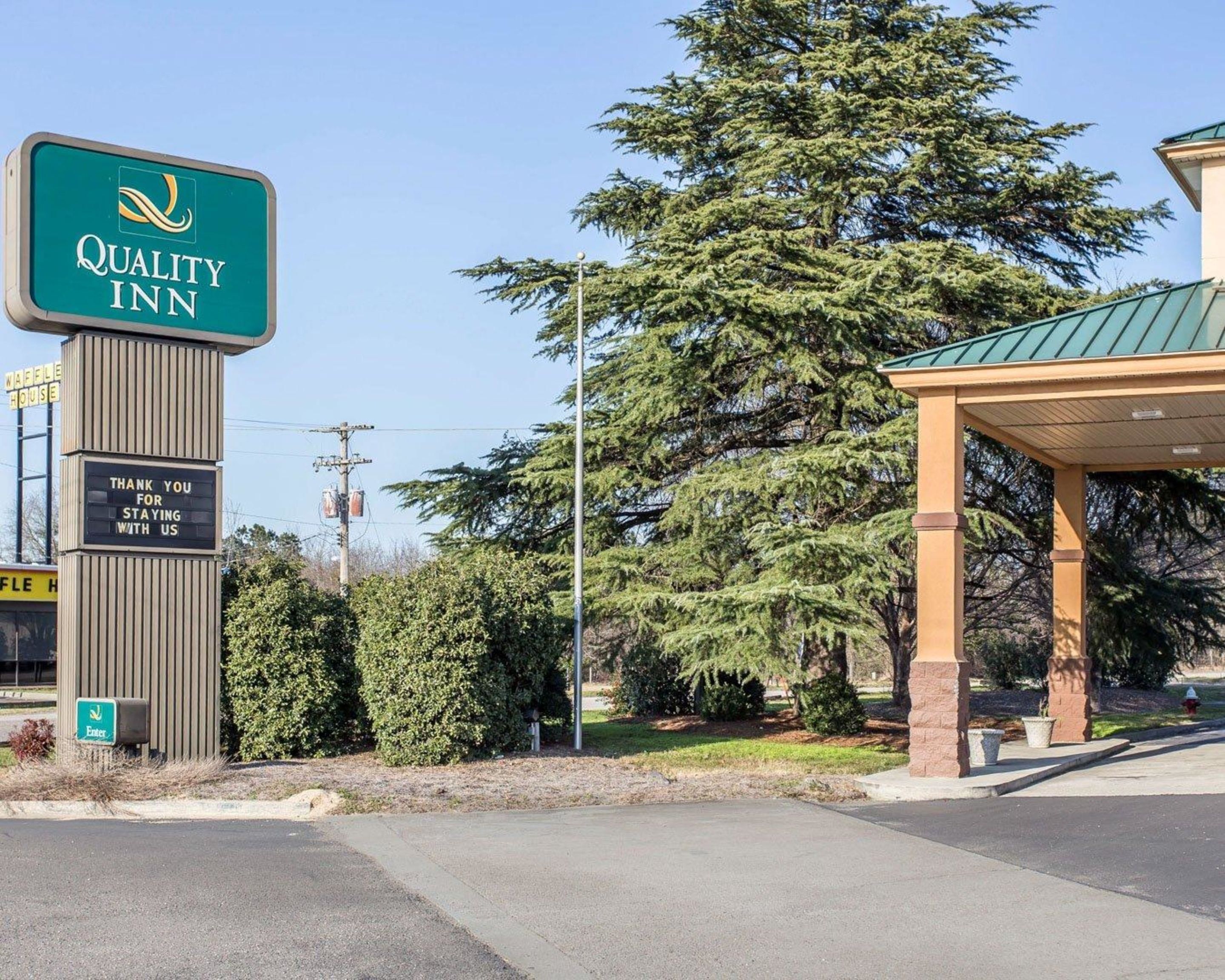 Señal de Quality Inn con un mensaje de agradecimiento, un área exterior cubierta y un árbol grande al fondo.
