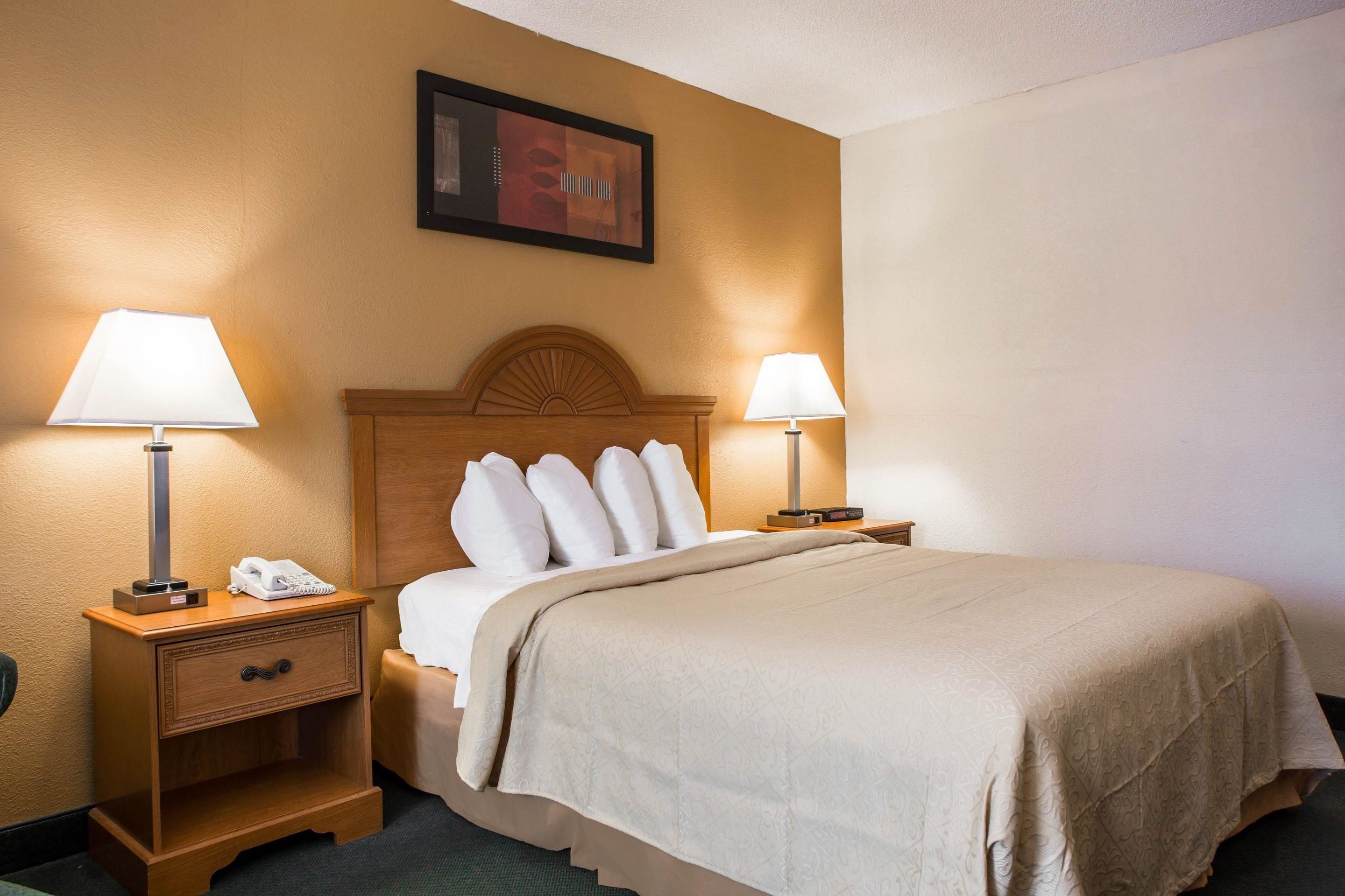 A hotel room with a bed, two bedside tables with lamps, a nightstand with a phone, and a framed picture on the wall.