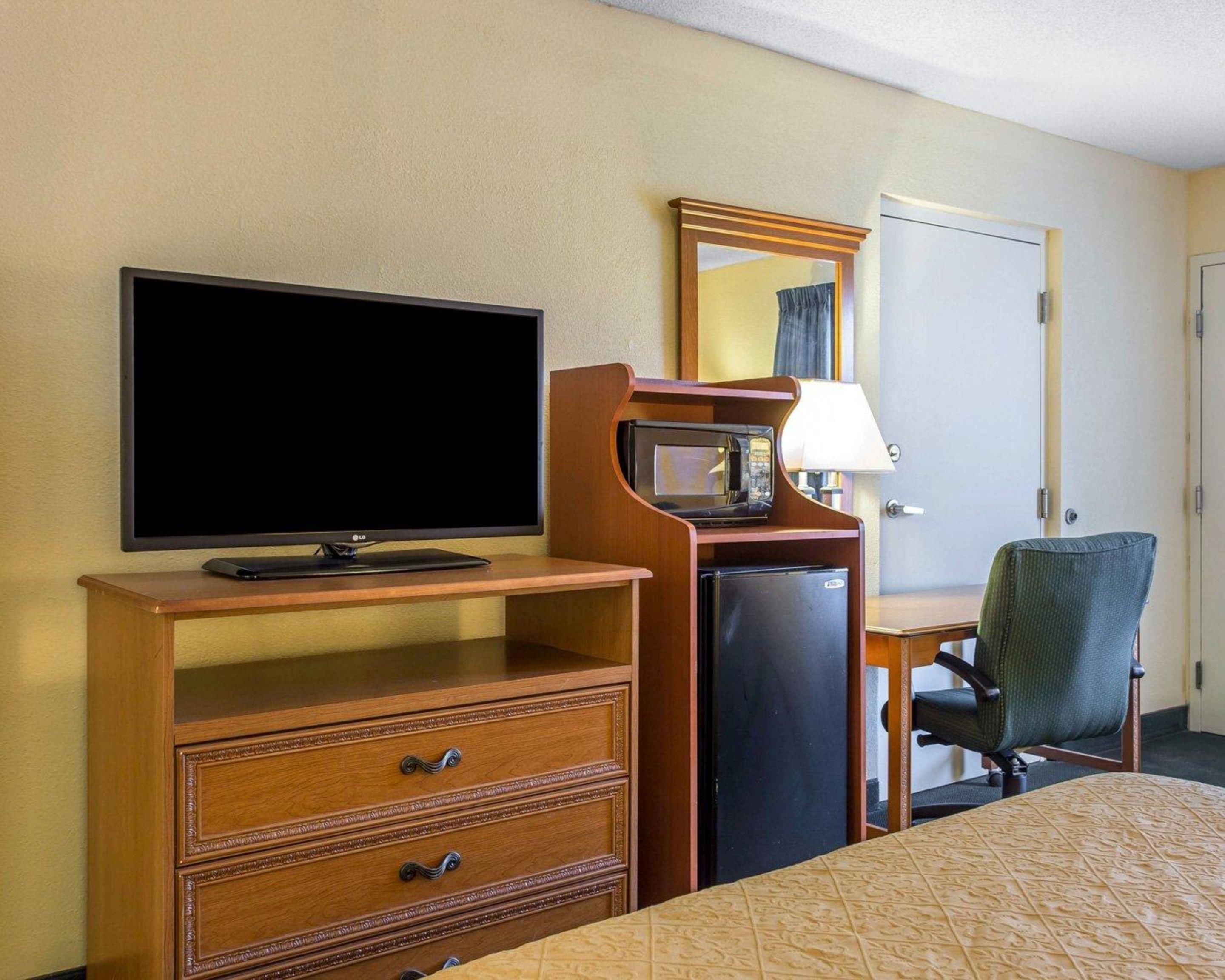 A hotel room with a flat-screen TV, a wooden dresser with drawers, a microwave, a small refrigerator, a desk with a lamp, a green chair, and a bed with a patterned bedspread.