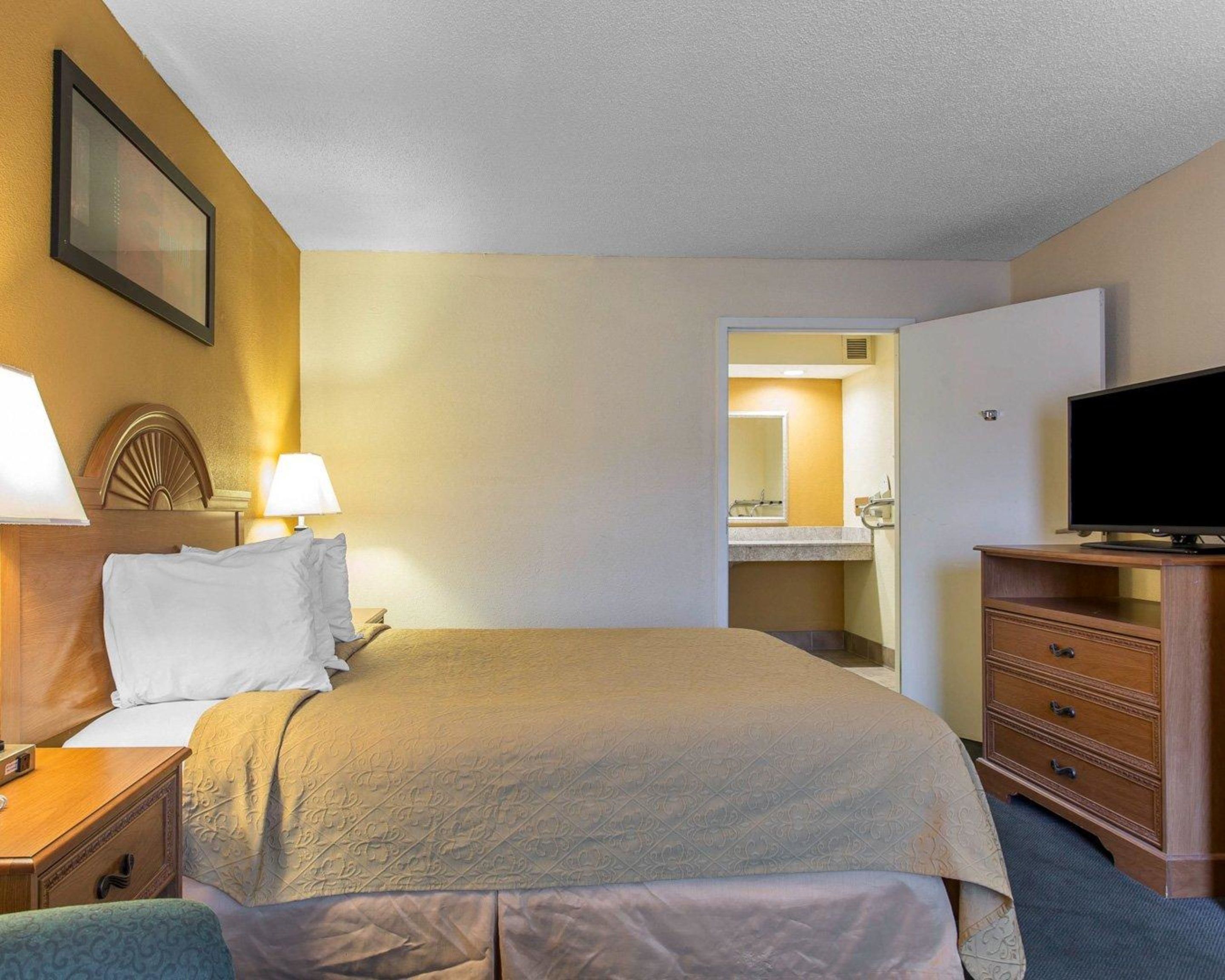 A hotel room with a bed, bedside table, lamp, TV, dresser, and a bathroom visible through an open door.