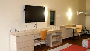 Une chambre d’hôtel équipée d’un grand téléviseur à écran plat fixé au mur, d’un bureau en bois avec des tiroirs, d’une chaise, d’une petite table avec des tasses et d’un miroir.