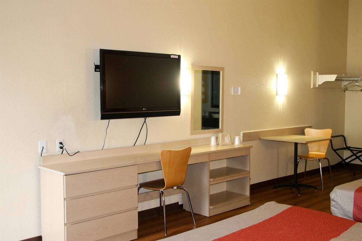 A hotel room with a large flat-screen TV mounted on the wall, a wooden desk with drawers, a chair, a small table with cups, and a mirror.