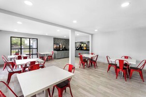 Un comedor moderno con mesas blancas y sillas rojas, una cocina al fondo y vistas a través de un ventanal.