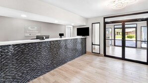 Un área de recepción con diseño moderno, que destaca por una pared de mosaicos, un mostrador de recepción, un televisor de pantalla plana y una puerta de vidrio que da al exterior.