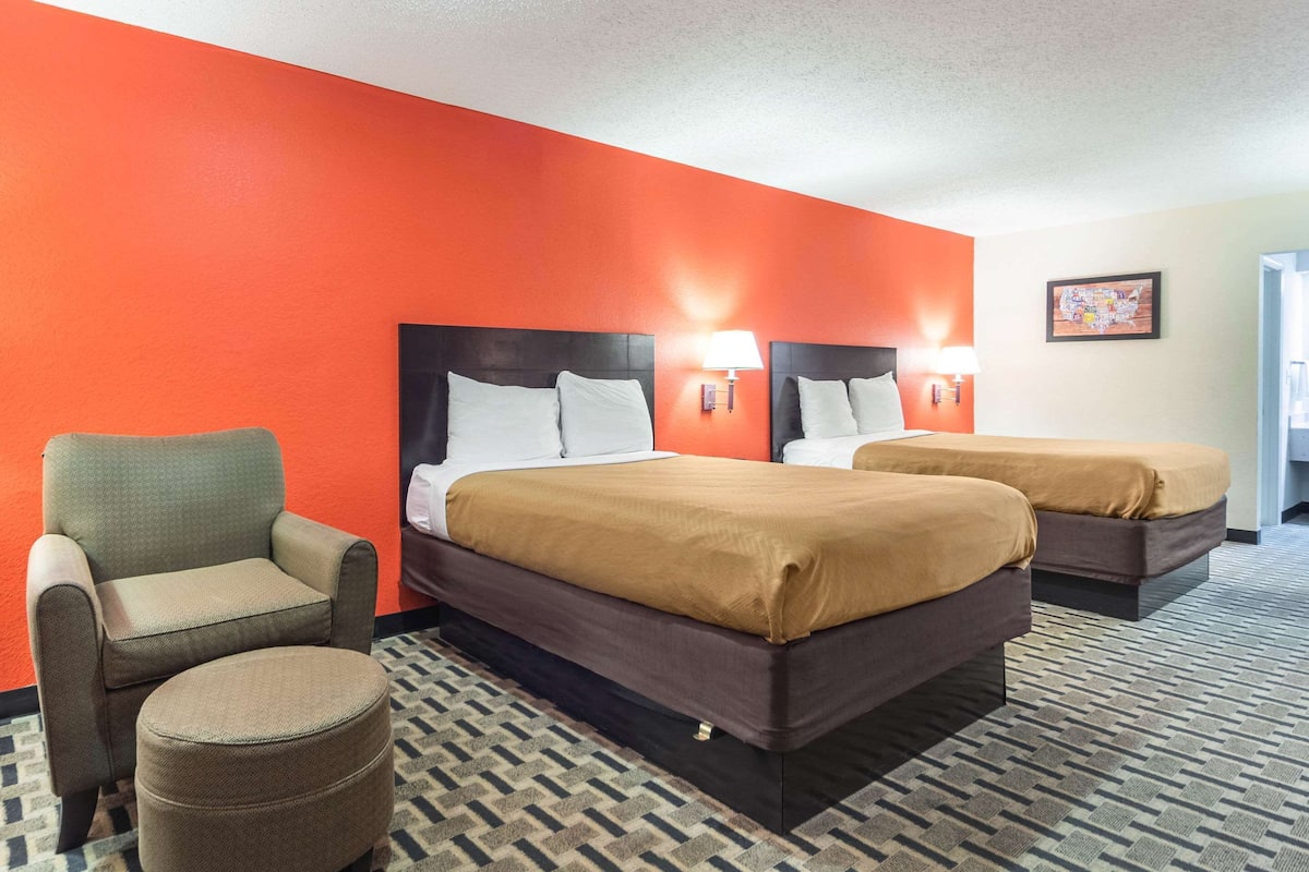 A hotel room with two beds, a chair, and a small ottoman.