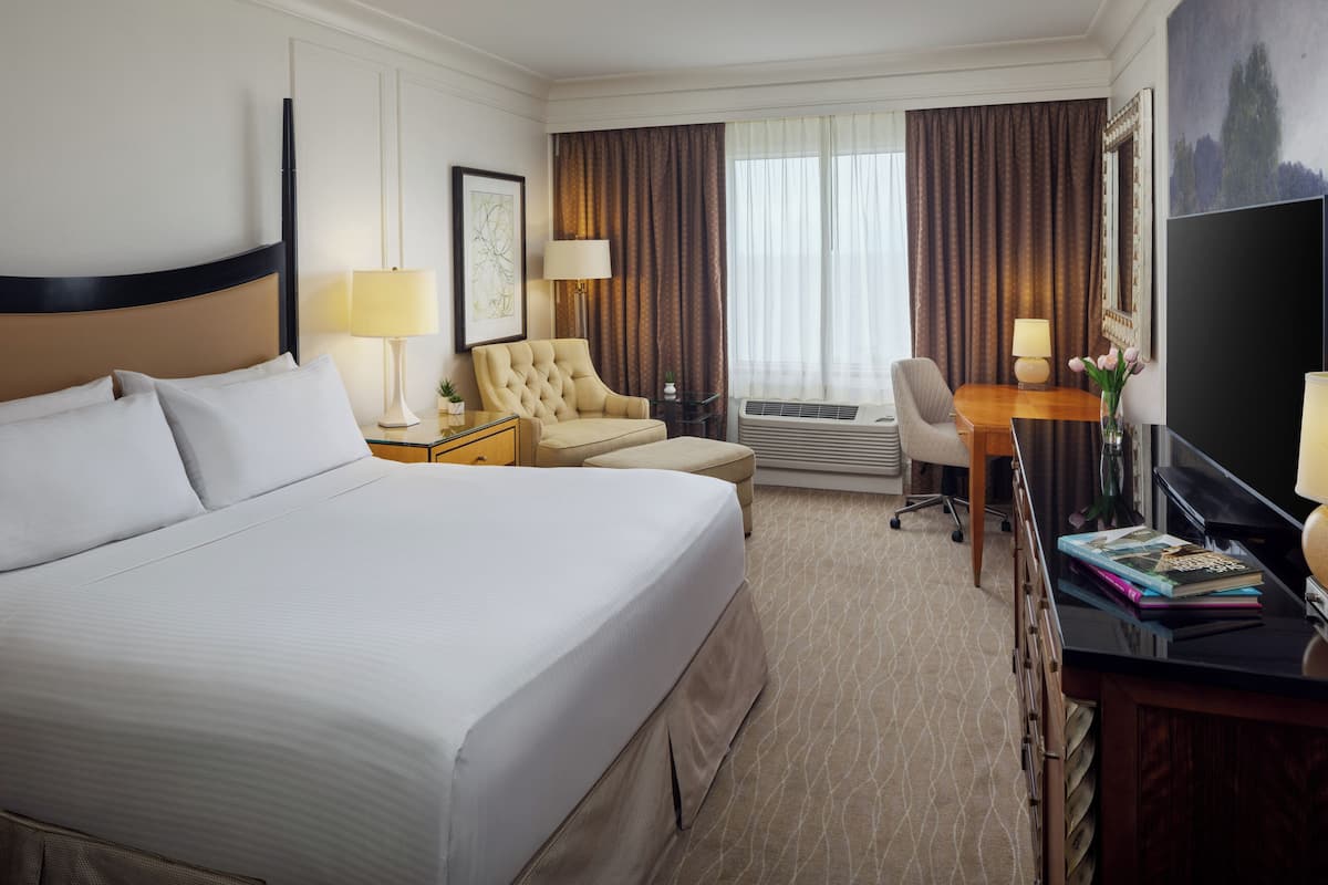A hotel room with a large bed, a desk with a computer, a chair, a TV, and a window with curtains.