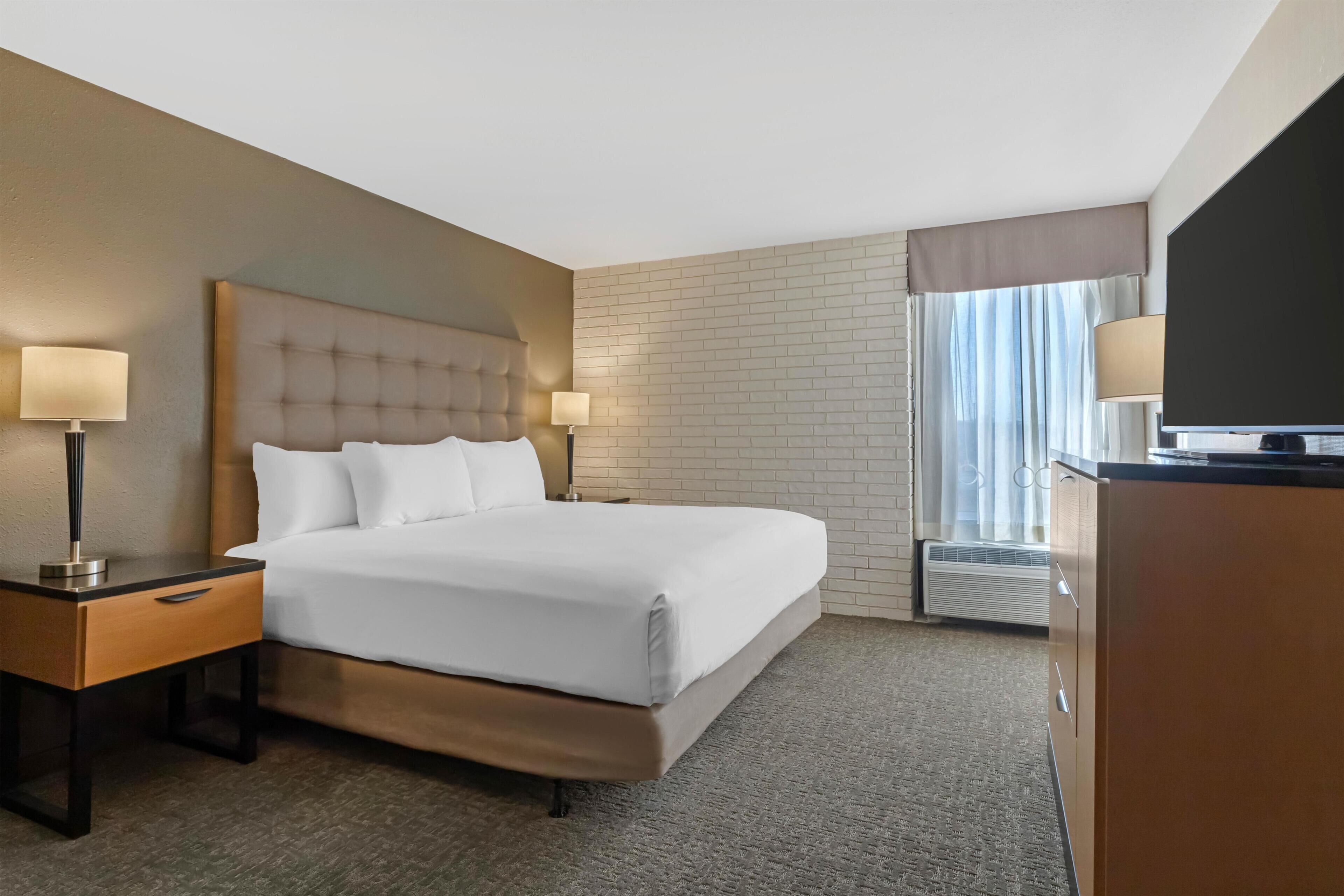 Une chambre d’hôtel avec un grand lit, deux tables de chevet avec des lampes, un téléviseur à écran plat, une commode et une fenêtre avec des rideaux.