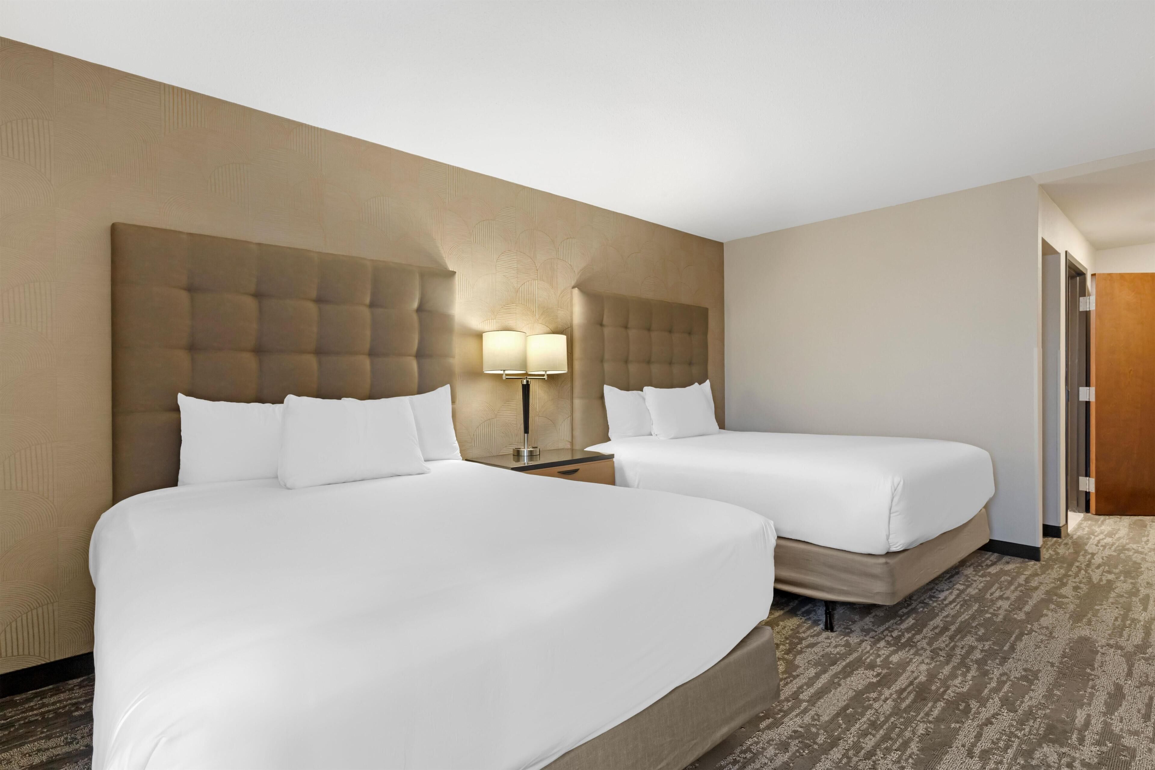 A hotel room with two beds, a large headboard, a nightstand with a lamp, and a carpeted floor.