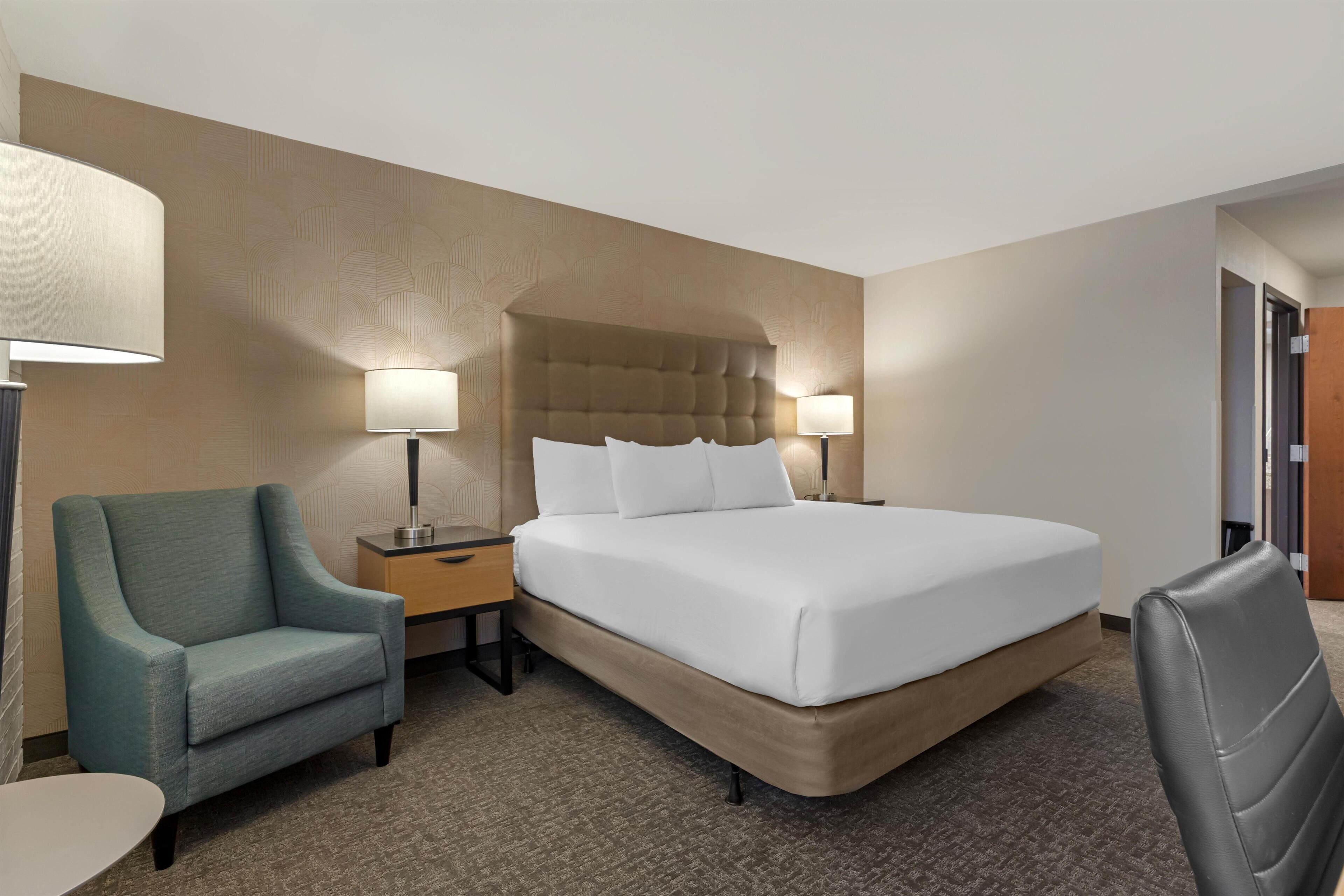 Habitación de hotel con una cama grande, dos lámparas de noche, una mesita pequeña, una silla y un buró.