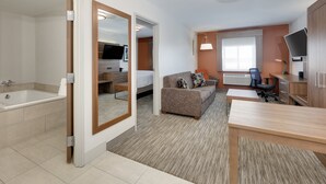 A modern hotel room with a bathroom, a living area with a sofa and TV, and a desk with a chair.