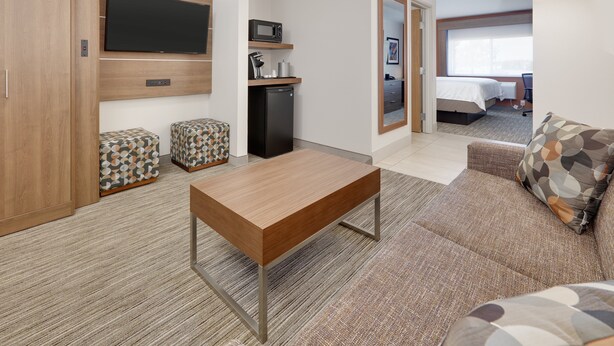 A hotel room with a wooden table, a sofa, a TV mounted on the wall, a microwave, and a small refrigerator.