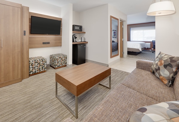 A hotel room with a wooden table, a sofa, a TV mounted on the wall, a microwave, and a small refrigerator.