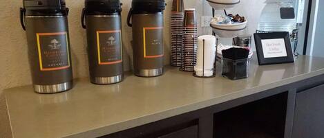 A buffet setup with hot water and coffee dispensers, stacked plates, and a sign for hot fresh coffee.