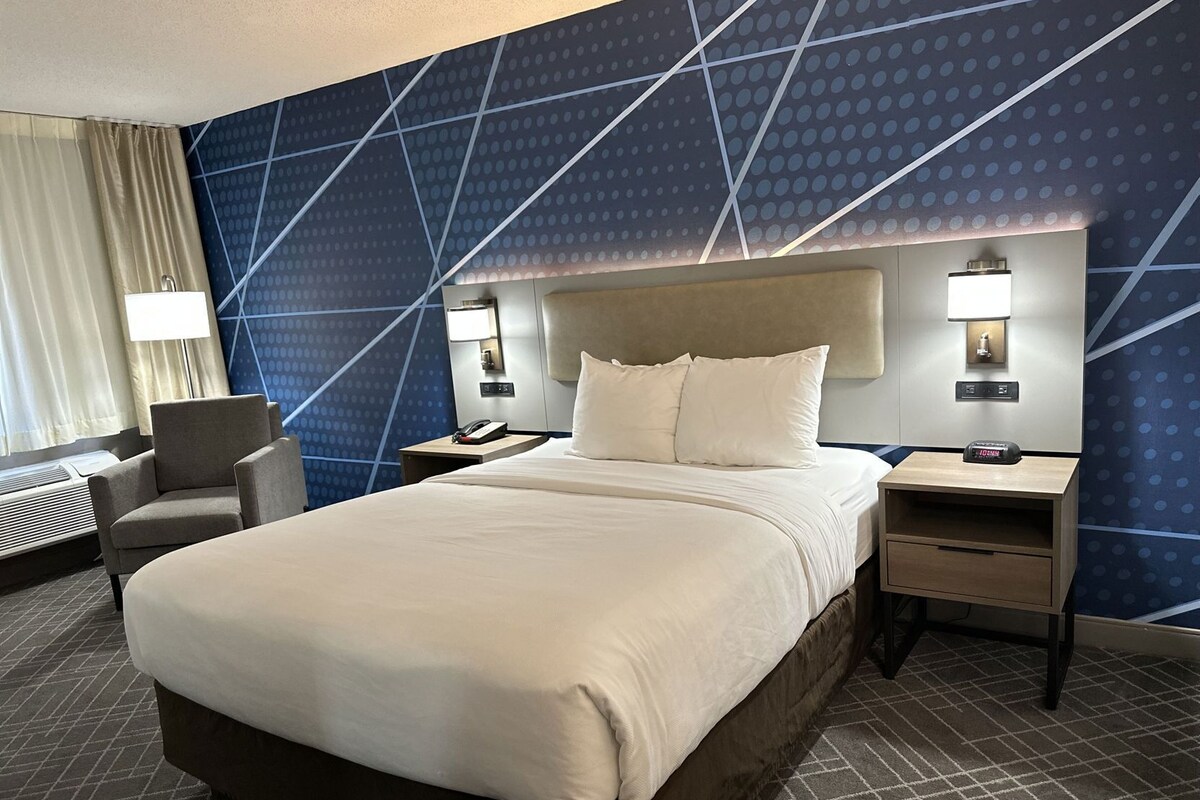 A hotel room with a large bed, a chair, a desk, a phone, and a wall clock.