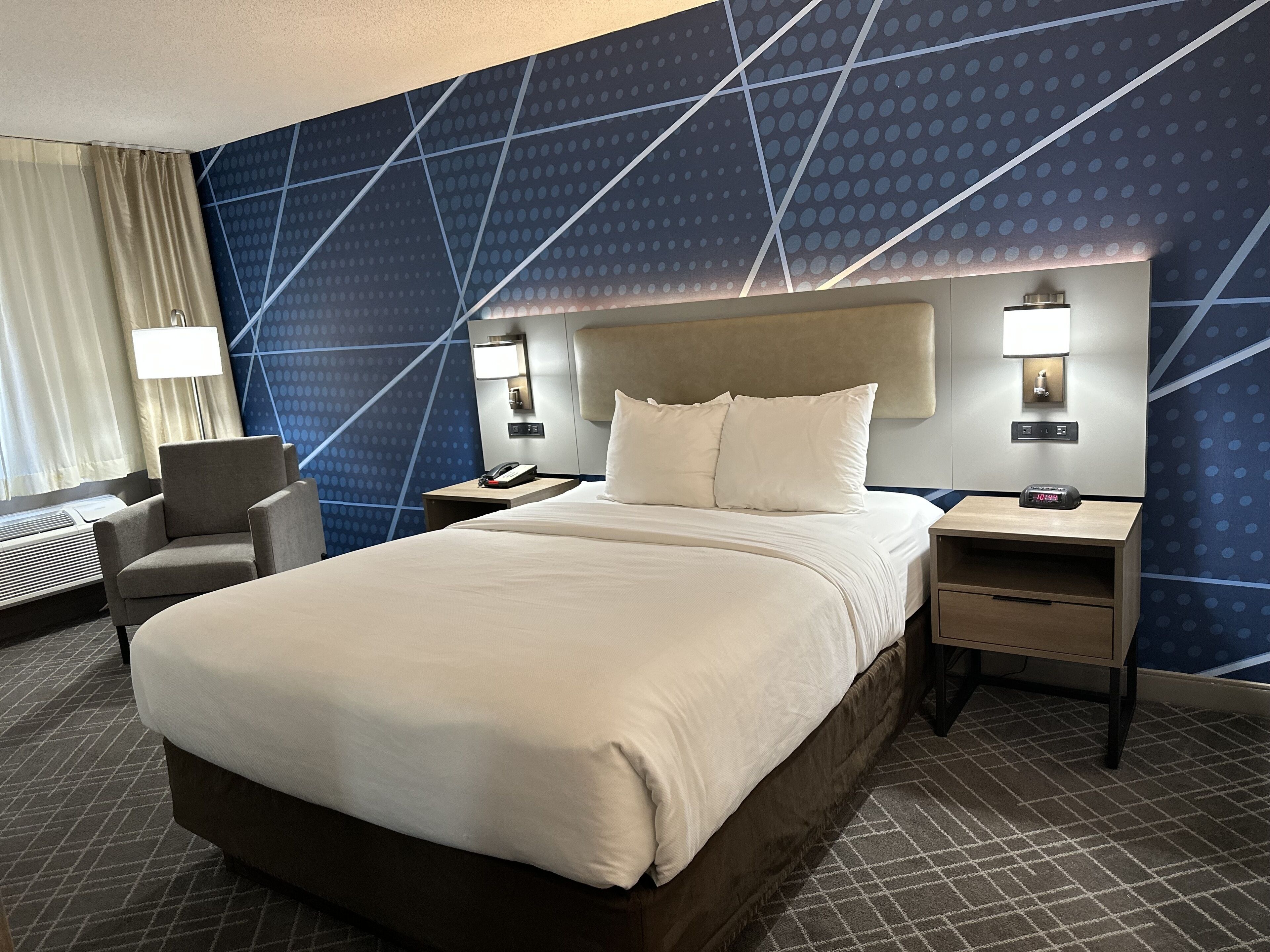 A hotel room with a large bed, a chair, a desk, and a wall with a geometric pattern.
