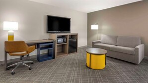 A modern hotel room with a flat-screen TV, a sofa, a desk with a chair, and a small round table.