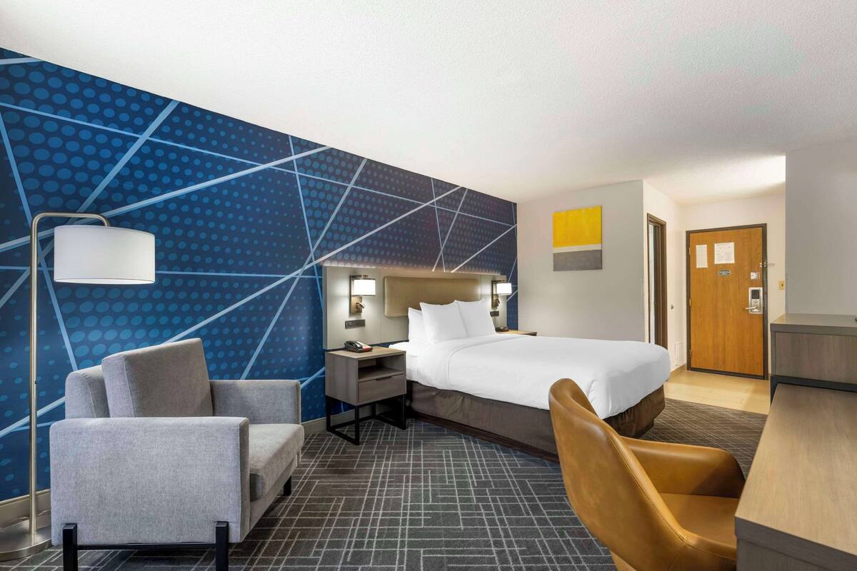 A hotel room with a bed, a desk, a chair, a lamp, and a wall with a geometric pattern.