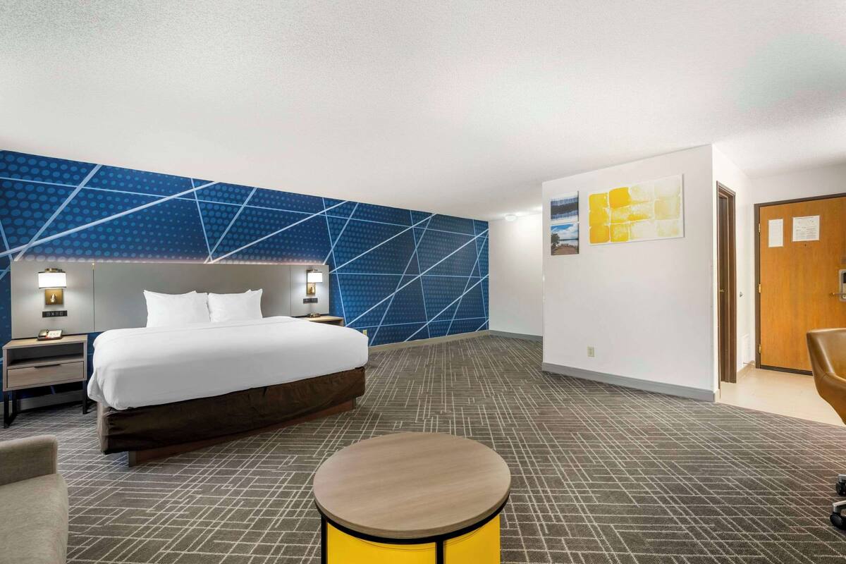 A hotel room with a large bed, a round wooden table, a nightstand, and a wall with geometric patterns.