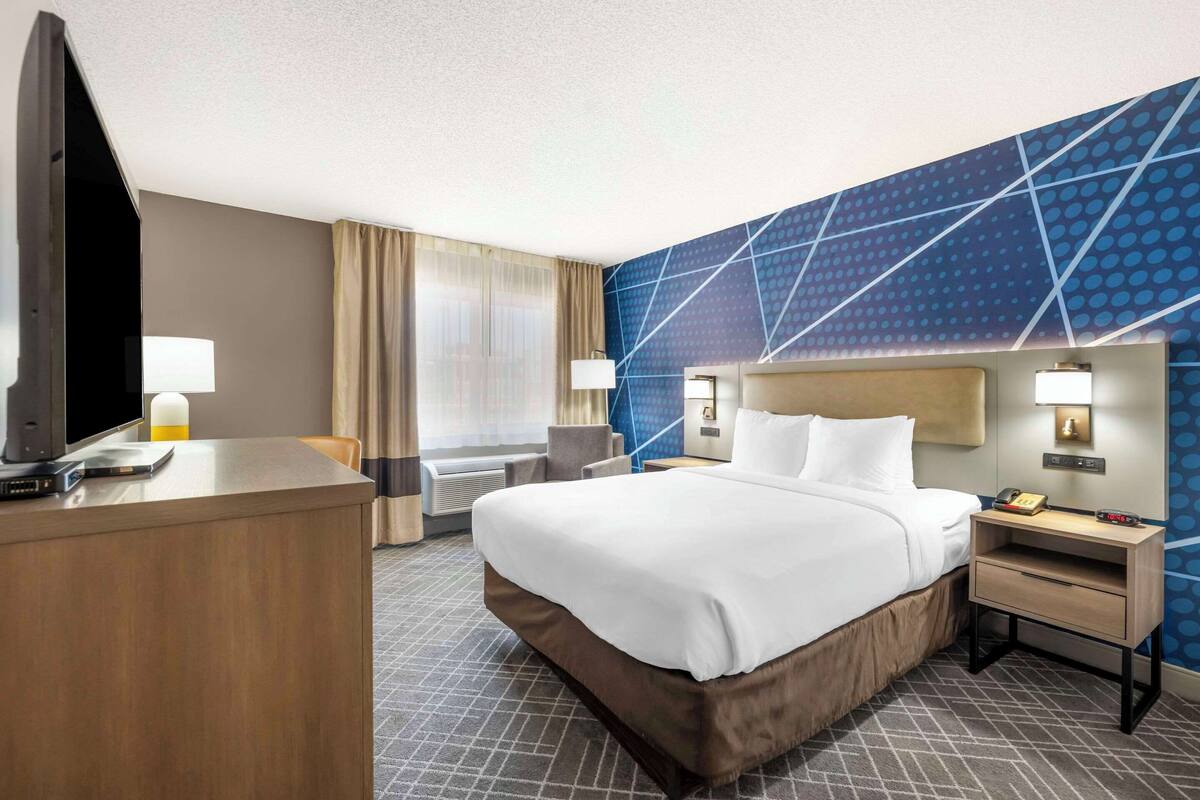 A hotel room with a large bed, a desk with a computer, a chair, and a nightstand with a lamp.
