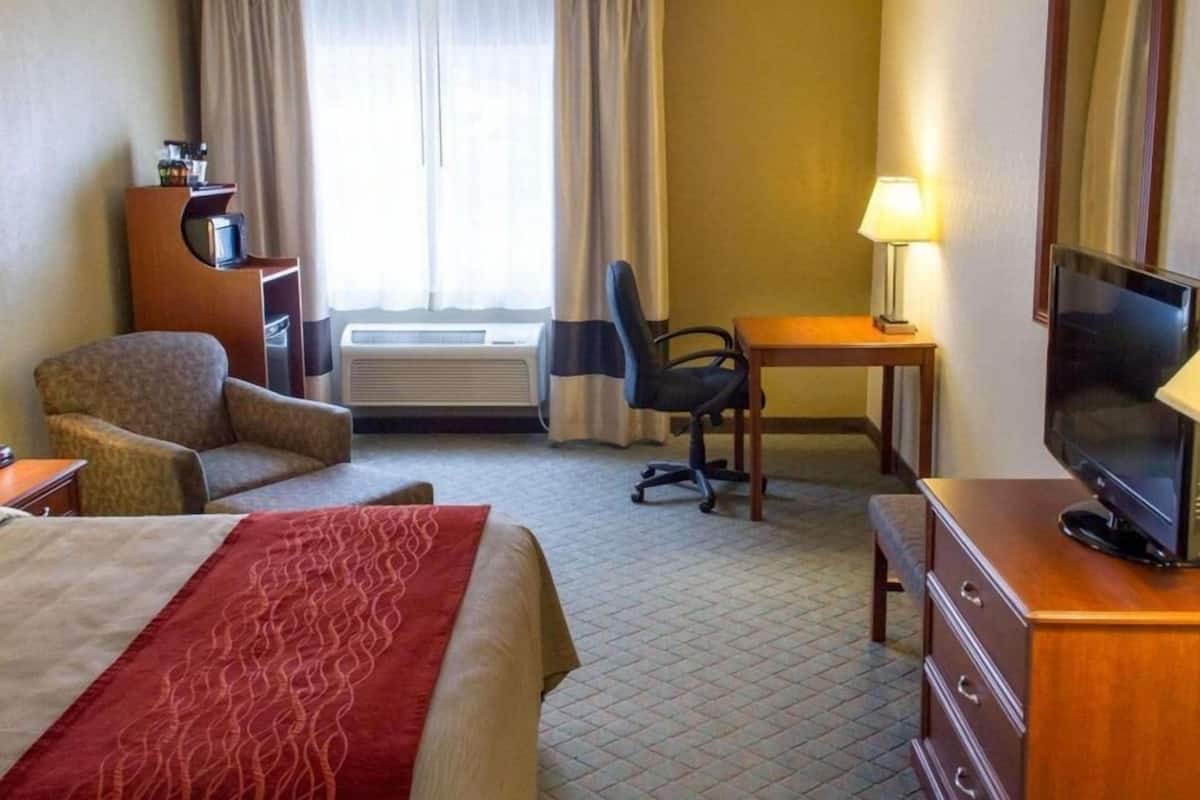 A hotel room with a bed, a desk, a chair, a television, an air conditioning unit, and a lamp.