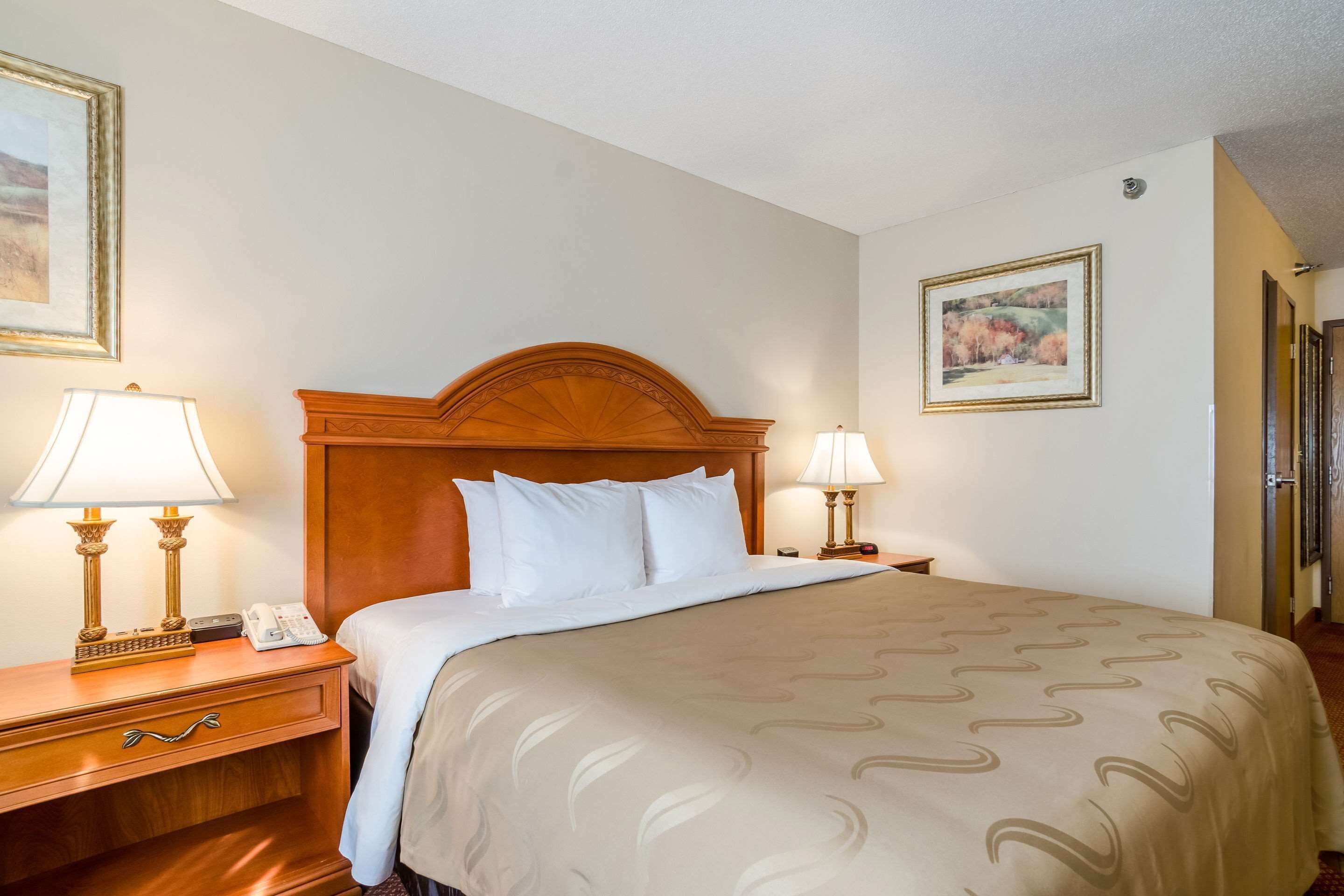 A hotel room with a wooden headboard, a large bed with white bedding, two bedside lamps, a nightstand with a telephone, and a framed picture on the wall.