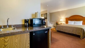A hotel room with a kitchenette including a microwave, coffee maker, and dishwasher. There is a bed with a wooden headboard and a nightstand with a lamp.